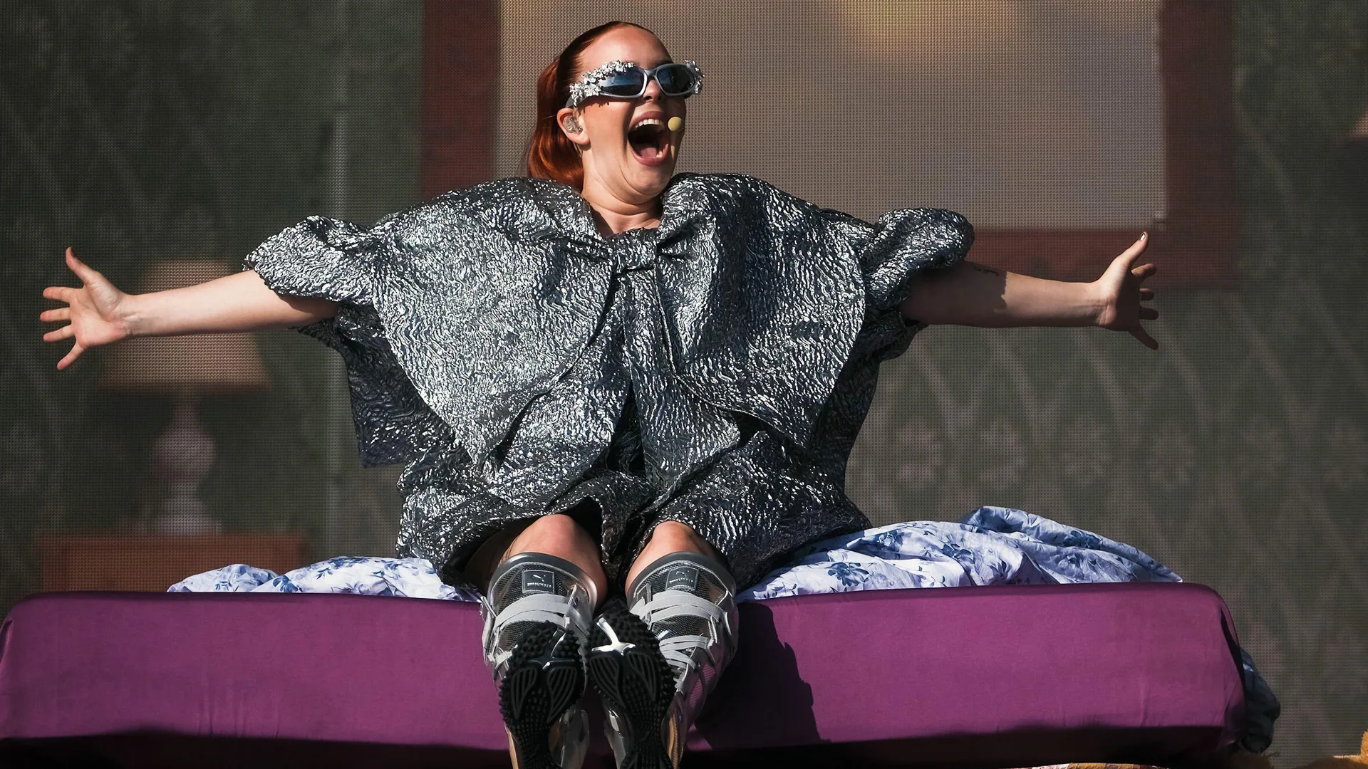 A photograph of the singer Anne-Marie performing at Glastonbury 2024 wearing a silver metallic dress with a giant bow on the front and metallic boots sat on a purple mattress with wallpaper behind her in a mock bedroom.