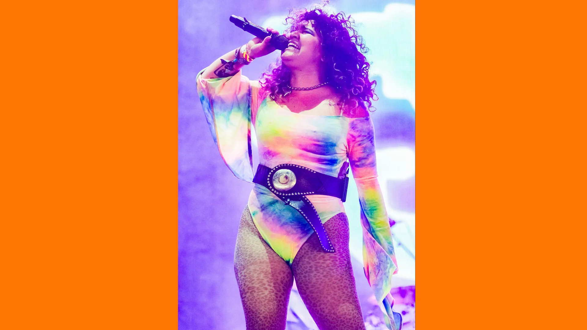 A photo of the singer Remi Wolf performing at Glastonbury 2024 wearing a rainbow coloured bodysuit and leopard print tights with a purple light behind her and orange borders to the image