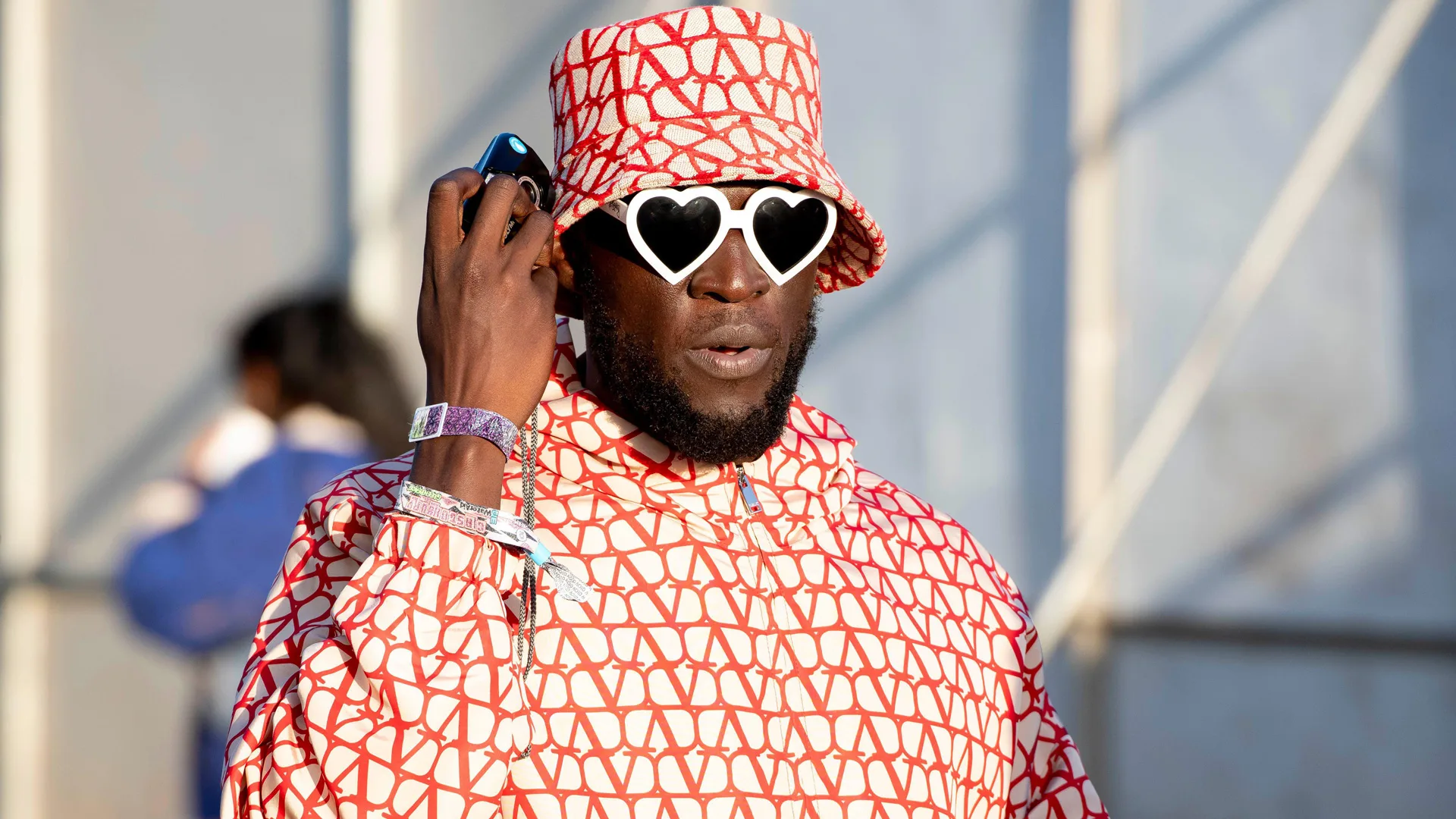 A photograph of the singer Stormzy at Glastonbury 2024 wearing a red and white Valentino raincoat and bucket hat with white heart shaped sunglasses