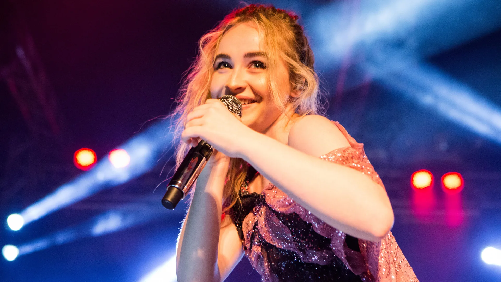 A photo of Sabrina Carpenter mid-performance smiling into the mic against a blue lit background