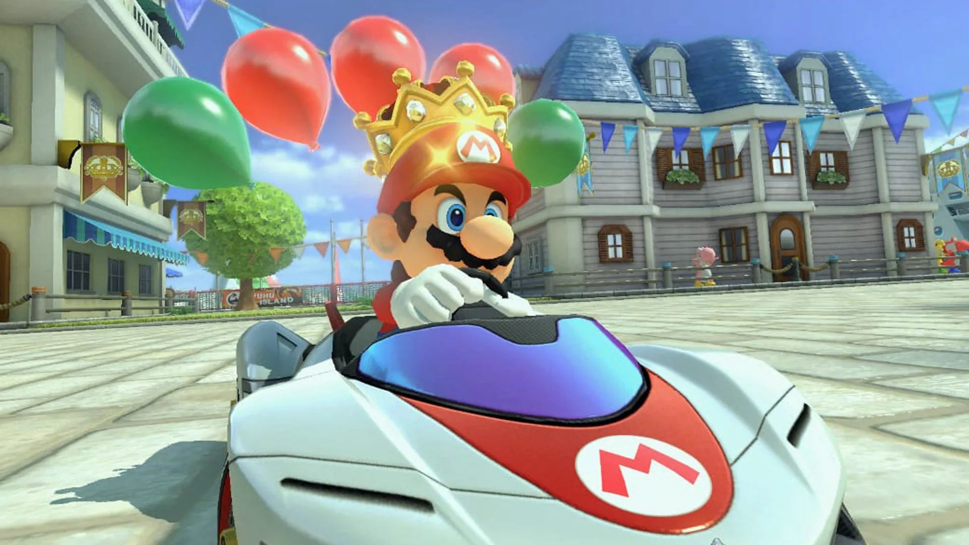 A still from Mario Kart showing Mario in his white and red car with the 'M' symbol on the bonnet driving through a town with balloons at the back of his vehicle