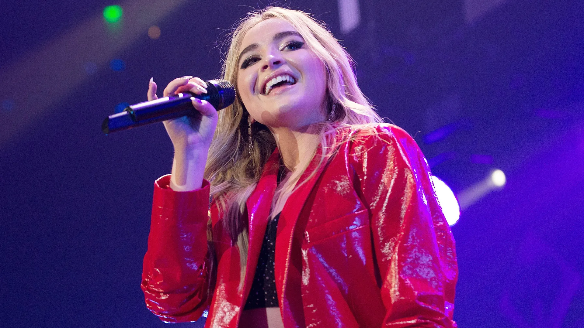 A photo of Sabrina Carpenter performing on stage in a red jacket with a mic to her face against a purple lit background
