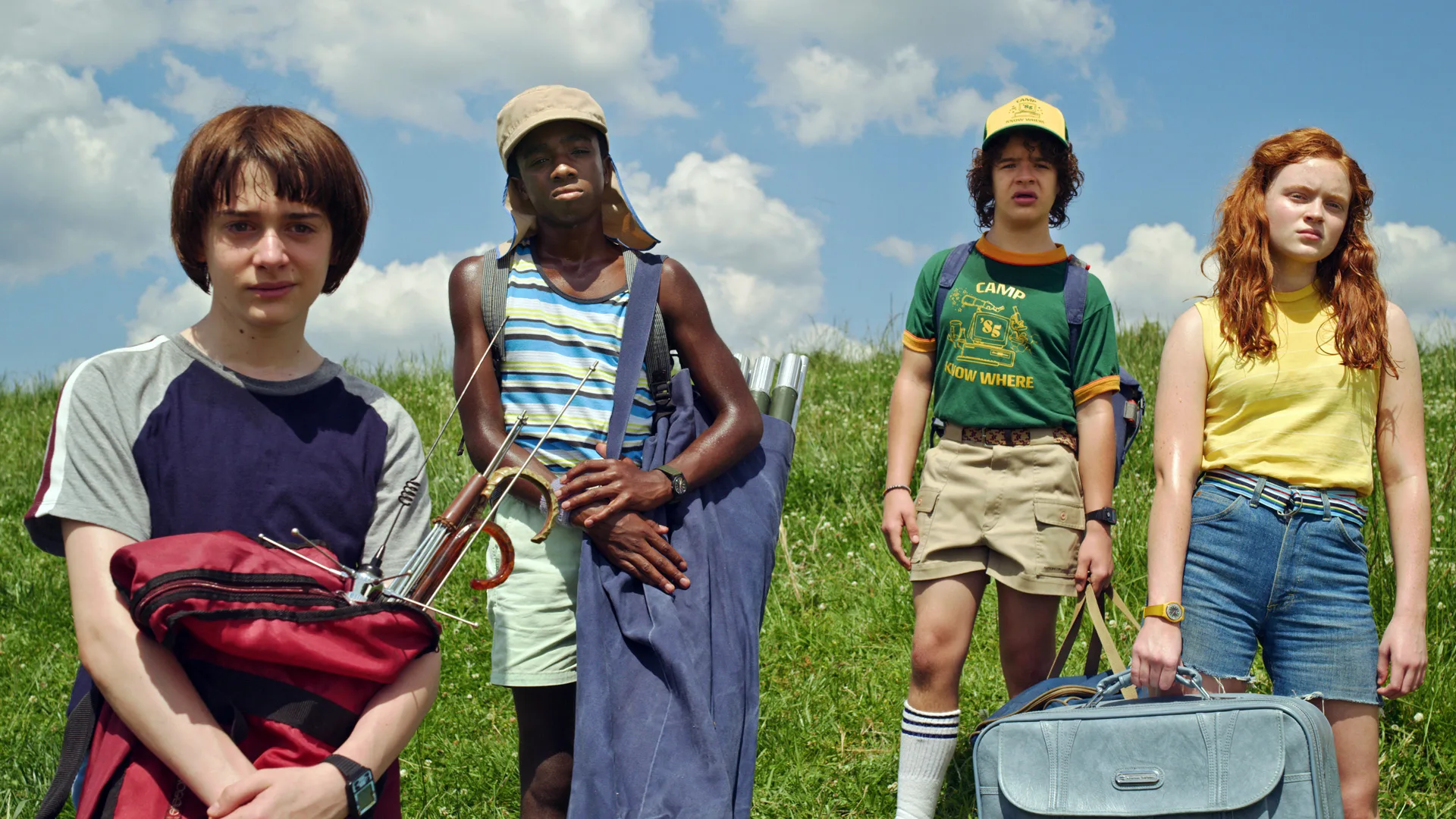 A photo of the characters Will, Dustin, Lucas and Max in Stranger Things all stood in a field in summer outfits against a blue cloudy sky