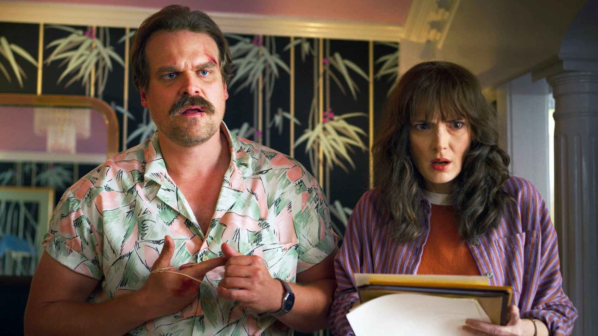 A photo of Stranger Things characters Hopper and Joyce stood with confused expressions on their face. Hopper is wearing a tropical shirt against a palm leaf picture on the wall.