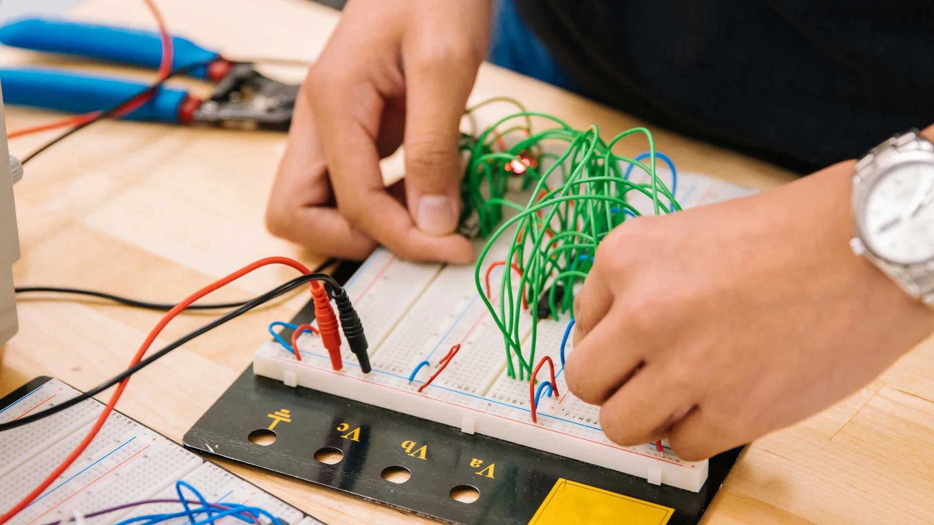 An image of two hands working on a circuit board of green and red wires on a table