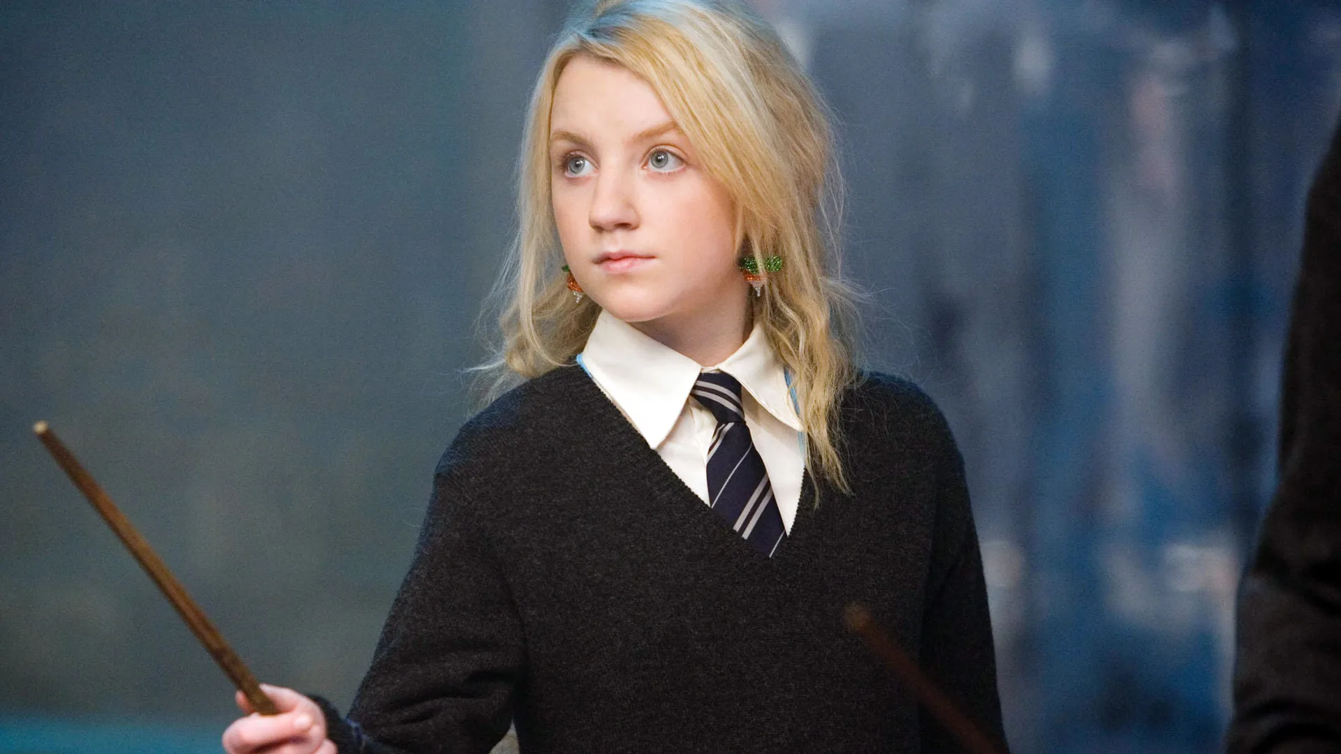 A photograph of Luna Lovegood from Harry Potter holding a wand in her Hogwarts uniform against a blue background