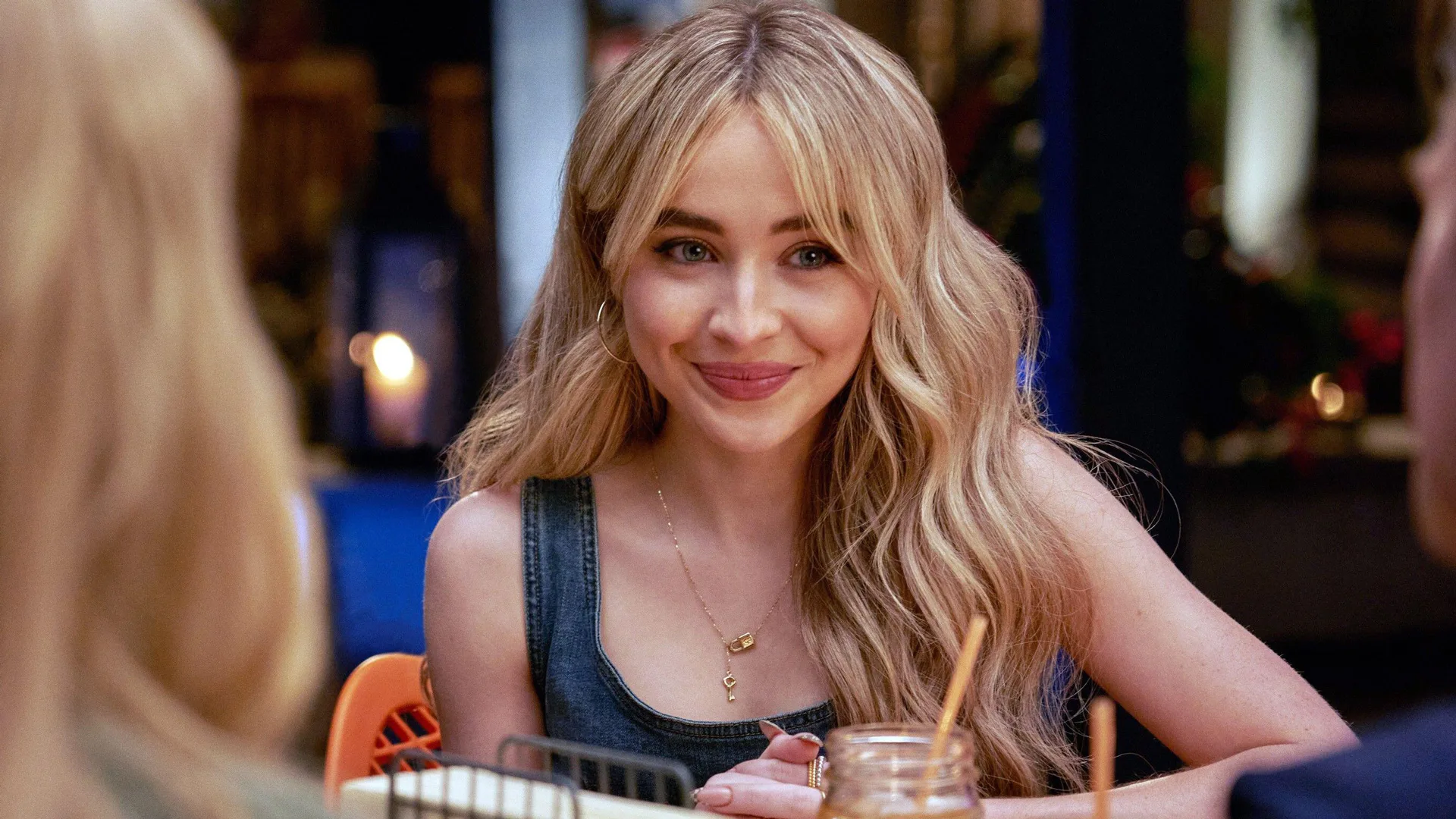A photograph of Sabrina Carpenter in the film Tall Girl smiling at a table with a black top
