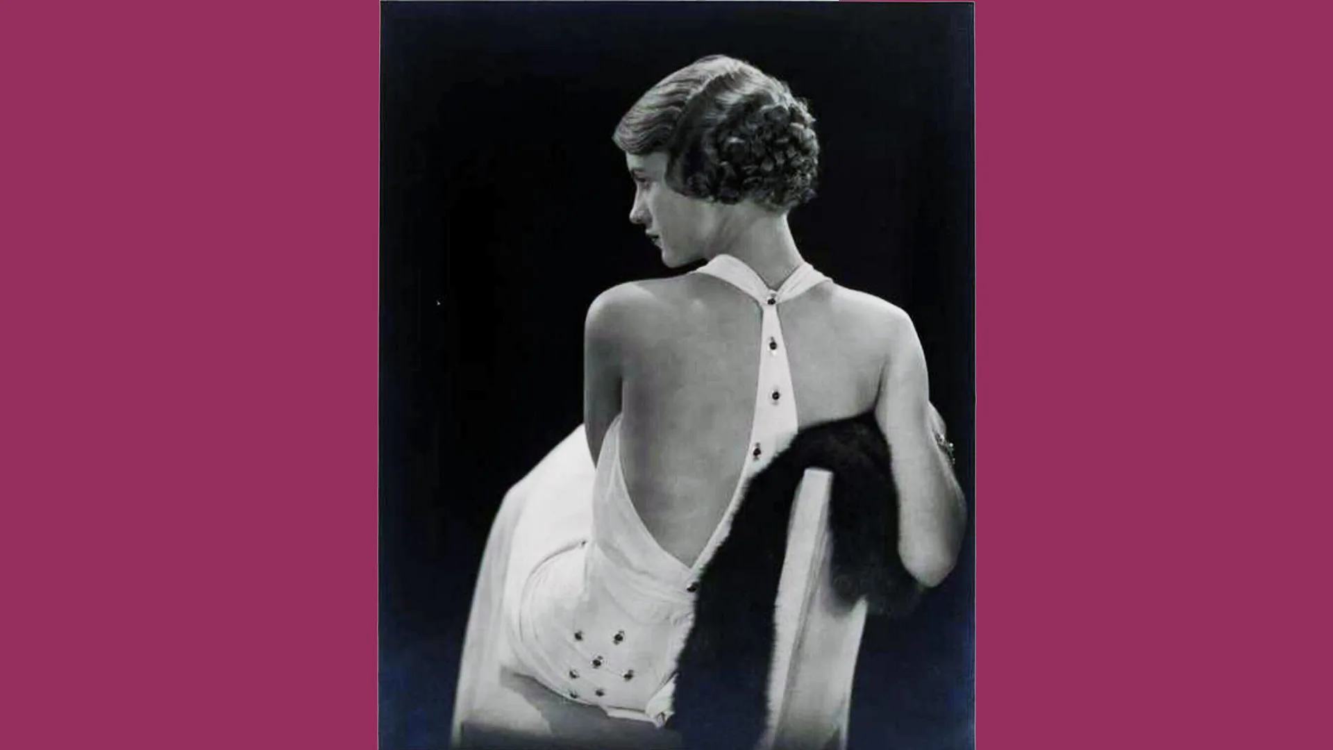 A black and white photograph of Lee Miller in a backless white dress sat on a chair with a fur coat underneath her arm