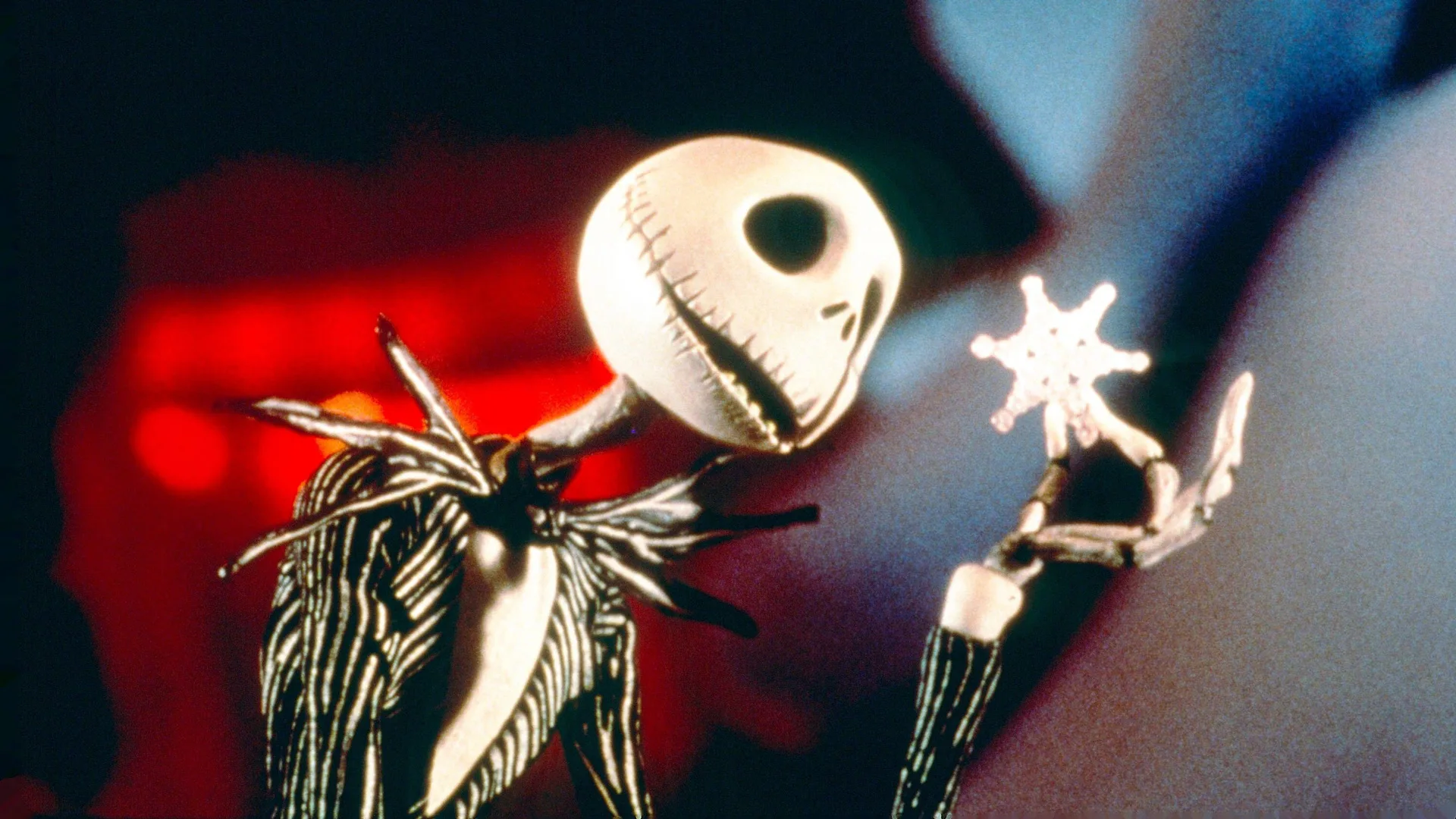 A still from the movie A Nightmare Before Christmas showing Jack Skellington holding a snowflake and inspecting it closely with a smile on his skull face against a winter lit up background