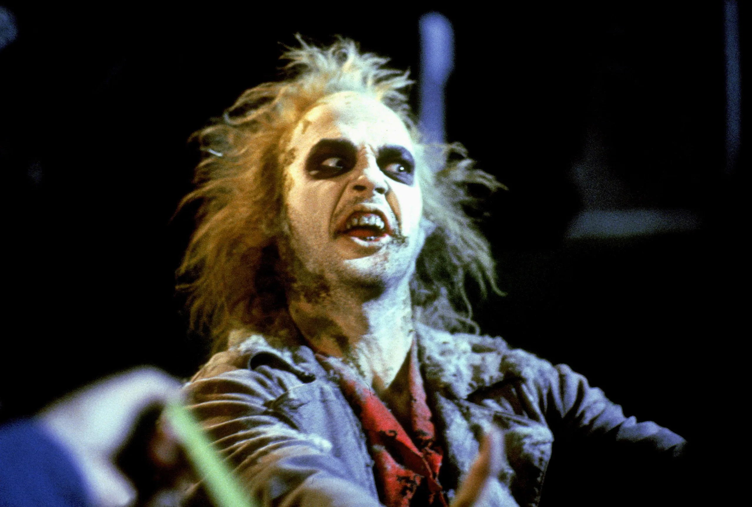 A still from the movie Beetlejuice (1988) showing the character Beetlejuice making a grimaced expression wearing his striped suit and shocked hair against a dark background