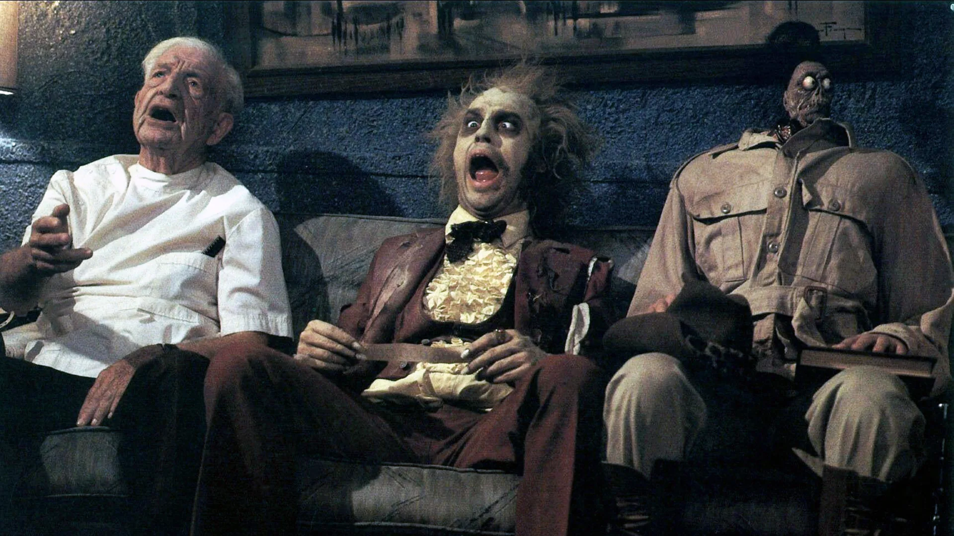 A still from the movie Beetlejuice showing the titular character sat on a sofa next to a shrunken headed man and an old man and they are screaming