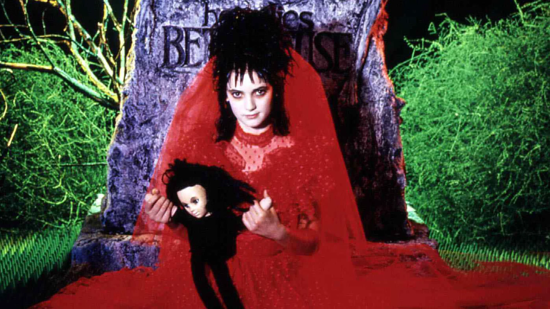 A promotional shot of Winona Ryder as the character Lydia from the movie Beetlejuice wearing a red bridal dress sat up against a gravestone holding a doll in black clothing with the background of a cemetarywith green hedges