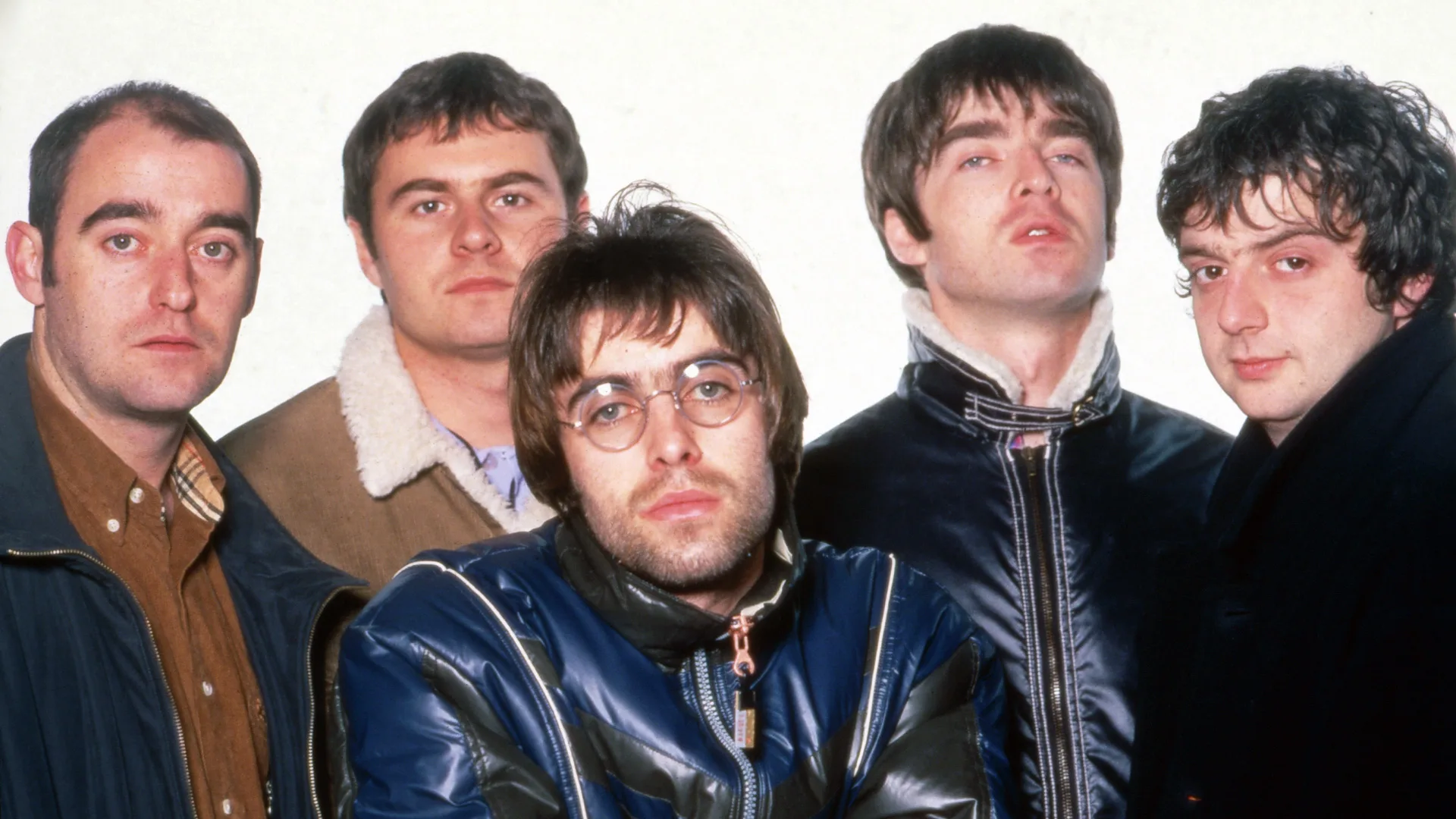 A photo of the band Oasis showing all 5 members looking at the camera with serious expressions against a white background