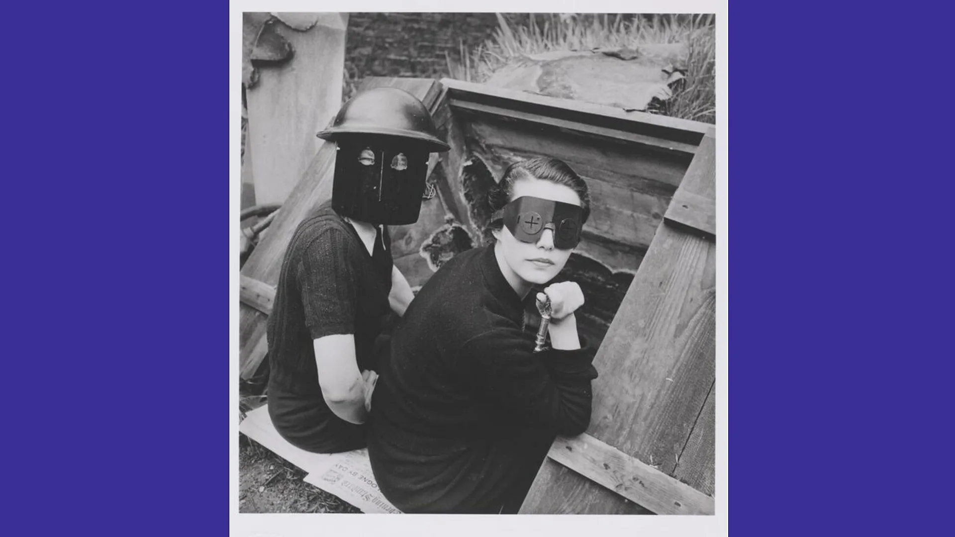 A black and white photograph of two women wearing masks as they hang over the edge of a bomb shelter during world war two, looking directly at the camera