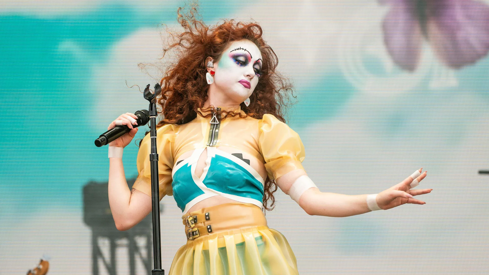 A photo of singer Chappell Roan performing on stage in a yellow and blue outfit wearing drag queen makeup and holding the mic against a painted blue sky cloudy background