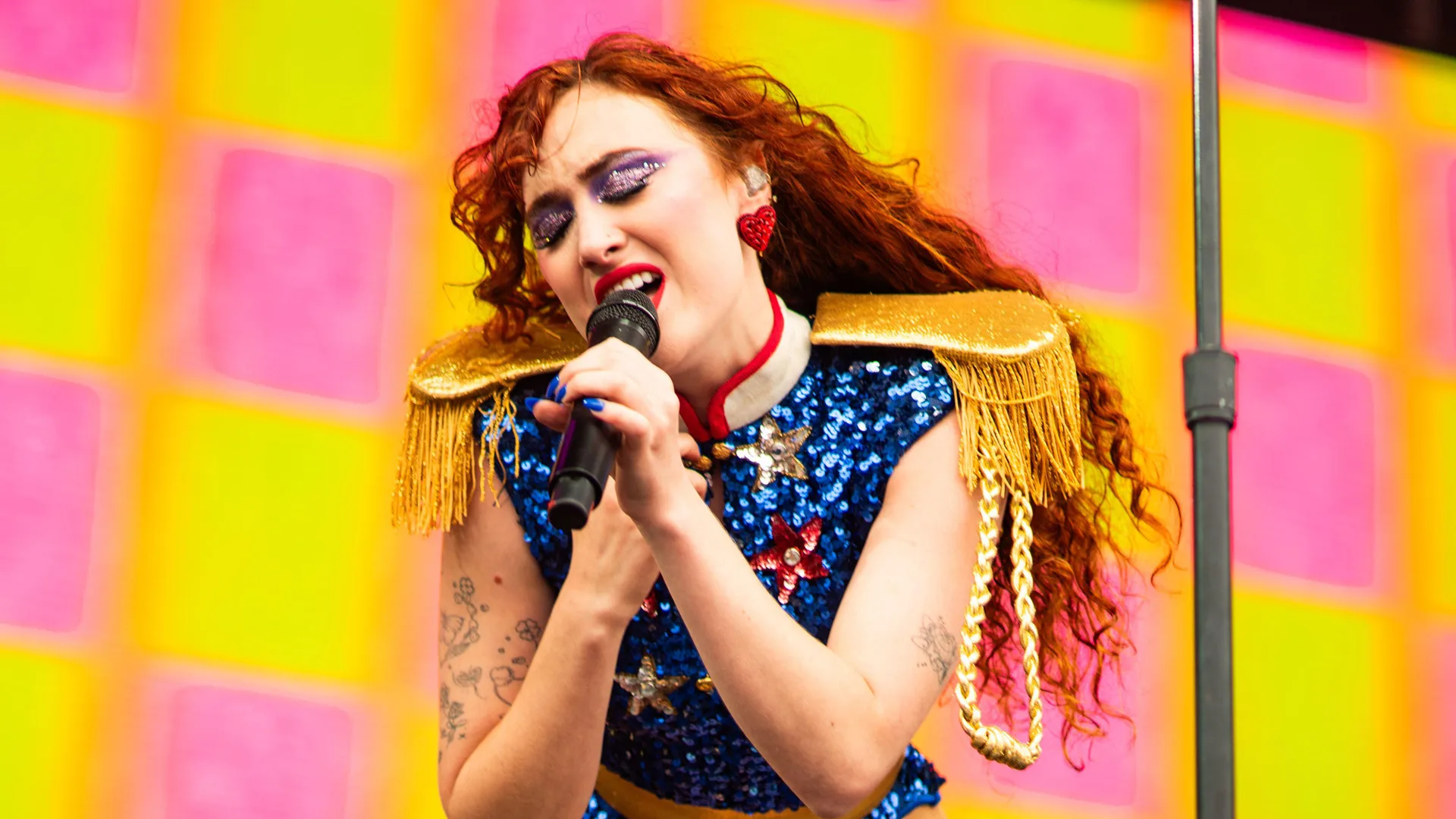 A photo of Chappell Roan singing on stage into the mic wearing a blue sparkly outfit with gold fringed shoulder pads against a bright yellow and pink squared background