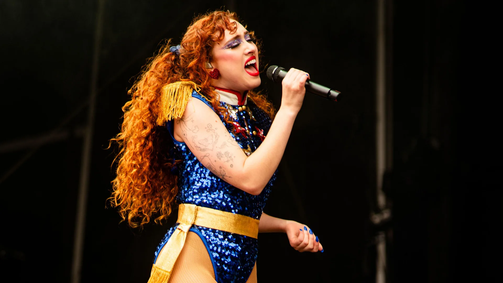 A photo of Chappell Roan singing on stage into a mic wearing a blue sparkly bodysuit with gold fringe shoulder pads against a black background