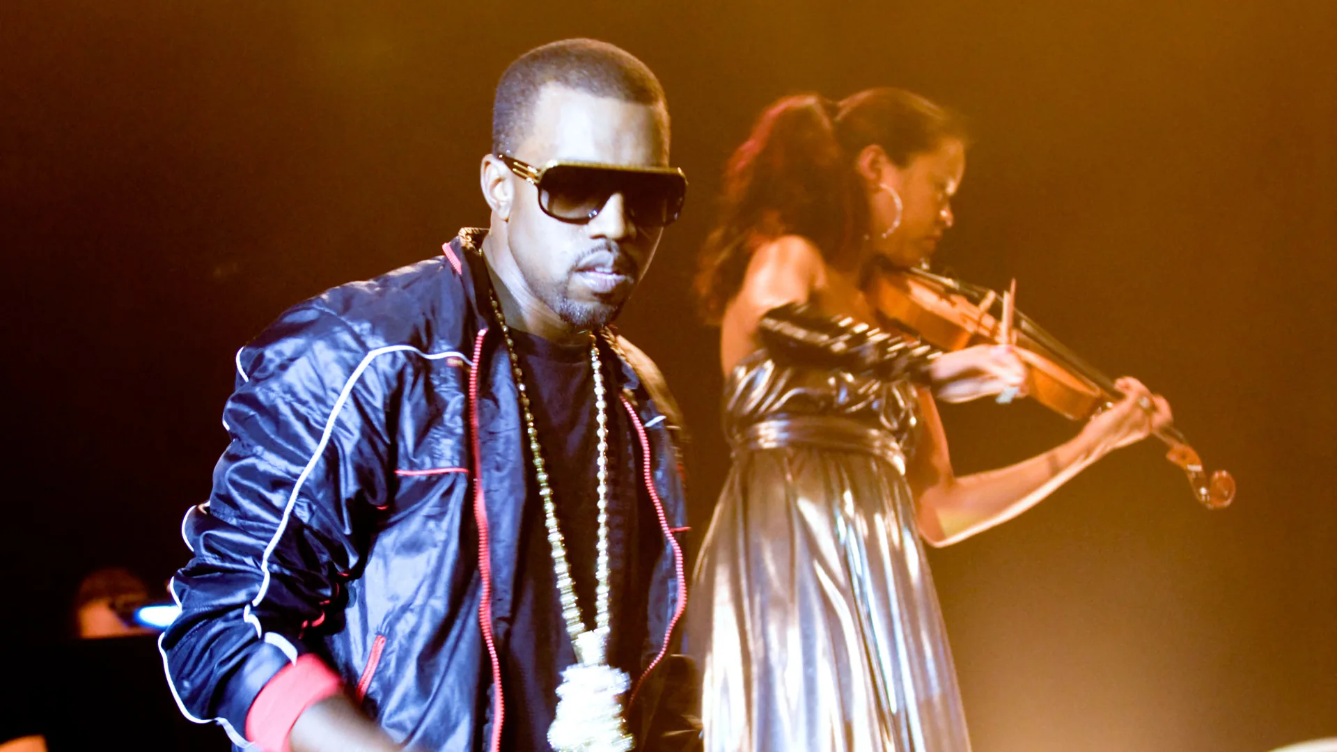 A photo of Kanye West performing on stage in a blue jacket next to a violinist.