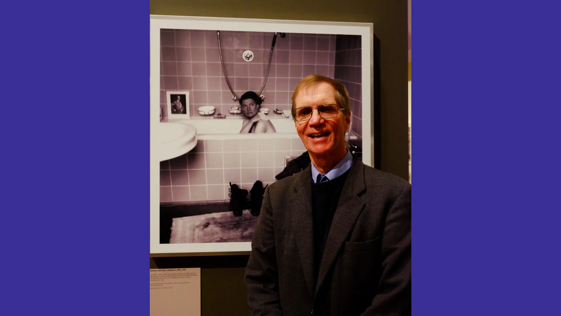 A photo of Lee Miller's son, Anthony Penrose stood smiling in front of one of her famous black and white photographs's of her sat inside Hitler's bathtub.