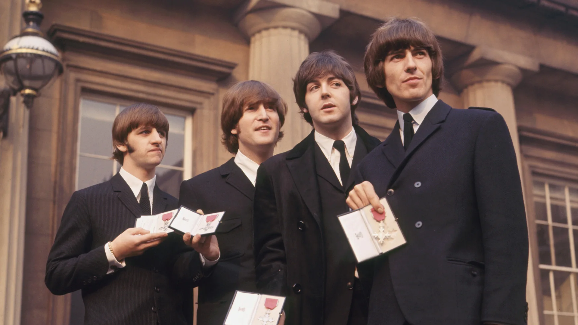 A photograph of The Beatles holding certificates stood outside a building. They are all wearing black and white suits.