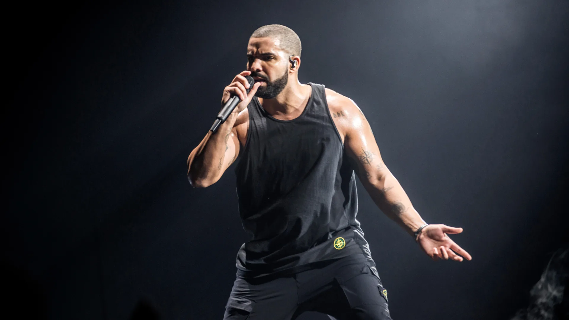 A photo of rapper Drake on stage singing into a mic wearing all black against a black background spotlit from the top.