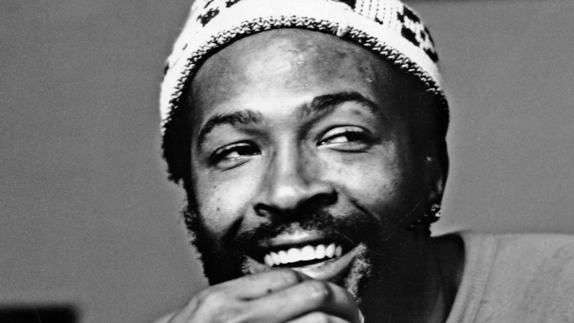 A black and white photograph of the singer Marvin Gaye holding his chin and smiling to the site. He is wearing a white beanie hat.