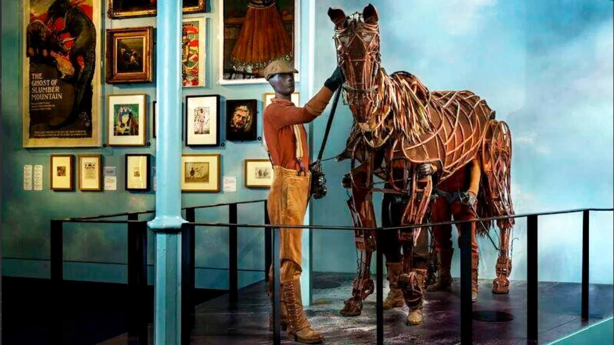 A photo of the lifesized puppet from the theatre show War Horse with a mannequin of a boy stood next to it for scale. The horse is in the Young V&A museum with pictures on the blue walls.