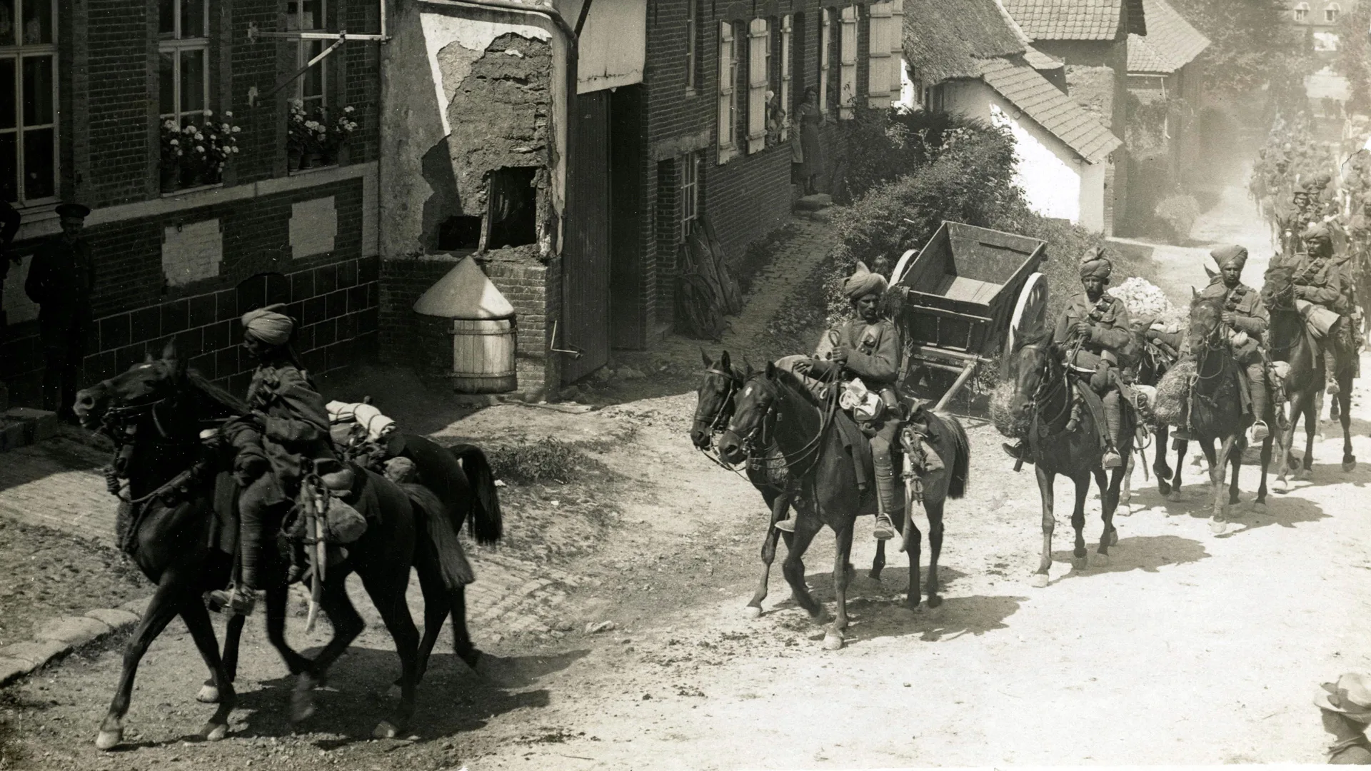 A black and white photo of a troop of Indian soldiers riding horses through a French village in WW1.