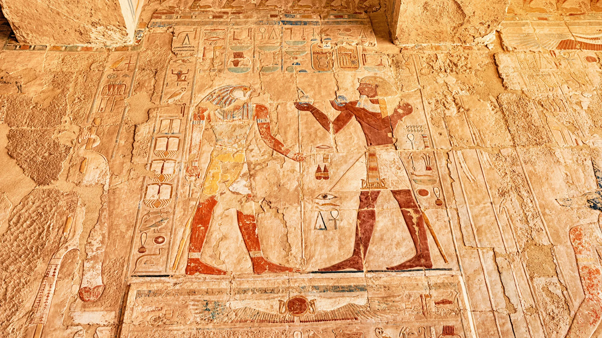 A photo of a wall with Ancient Egypt artwork depicting two Gods passing an organ to eachother surrounded by scales and other paraphenilia.