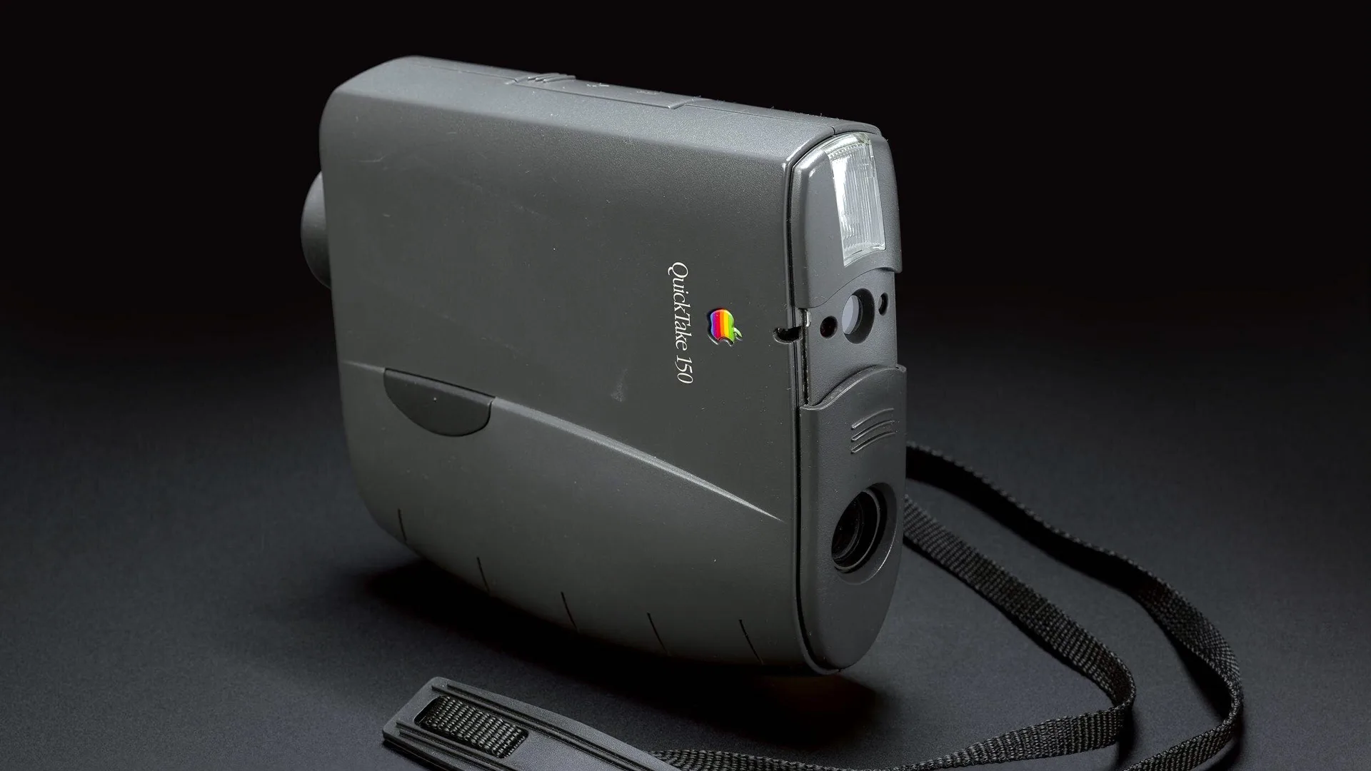 A photo of the first commercial digital camera made by Apple and Kodak showing a grey camera with a black strap against a black background.