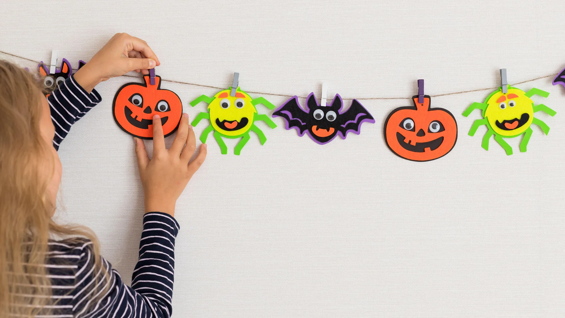 A photo of Halloween garland in the shapes of spiders, pumpkins, bats - being hung up by a girl in a striped top.