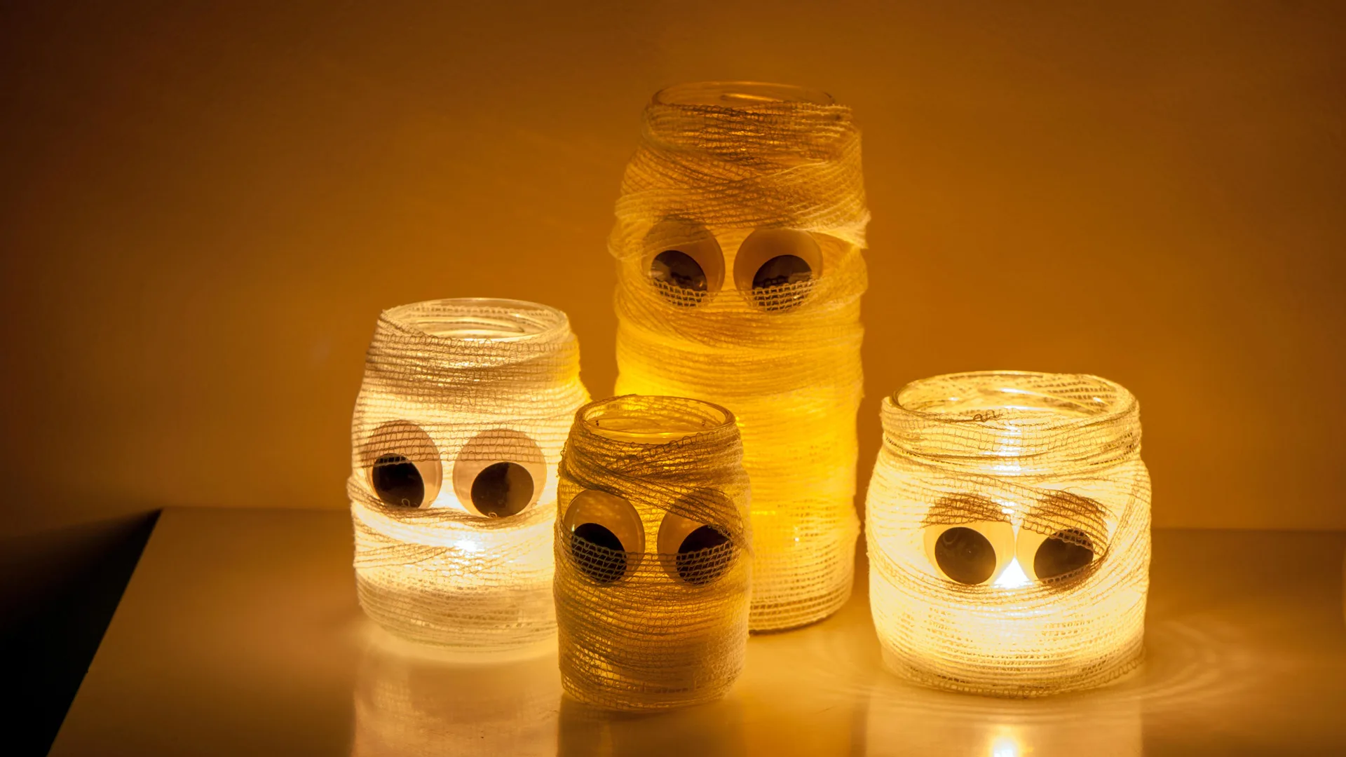 A photo of some Halloween jam jars decorated like Mummies with goog.y eyes and lights inside