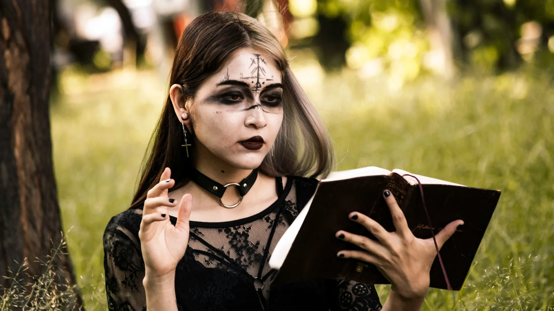 A photo of a woman holding a black book with dark gothic makeup stood in a forest with grass and trees behind her.