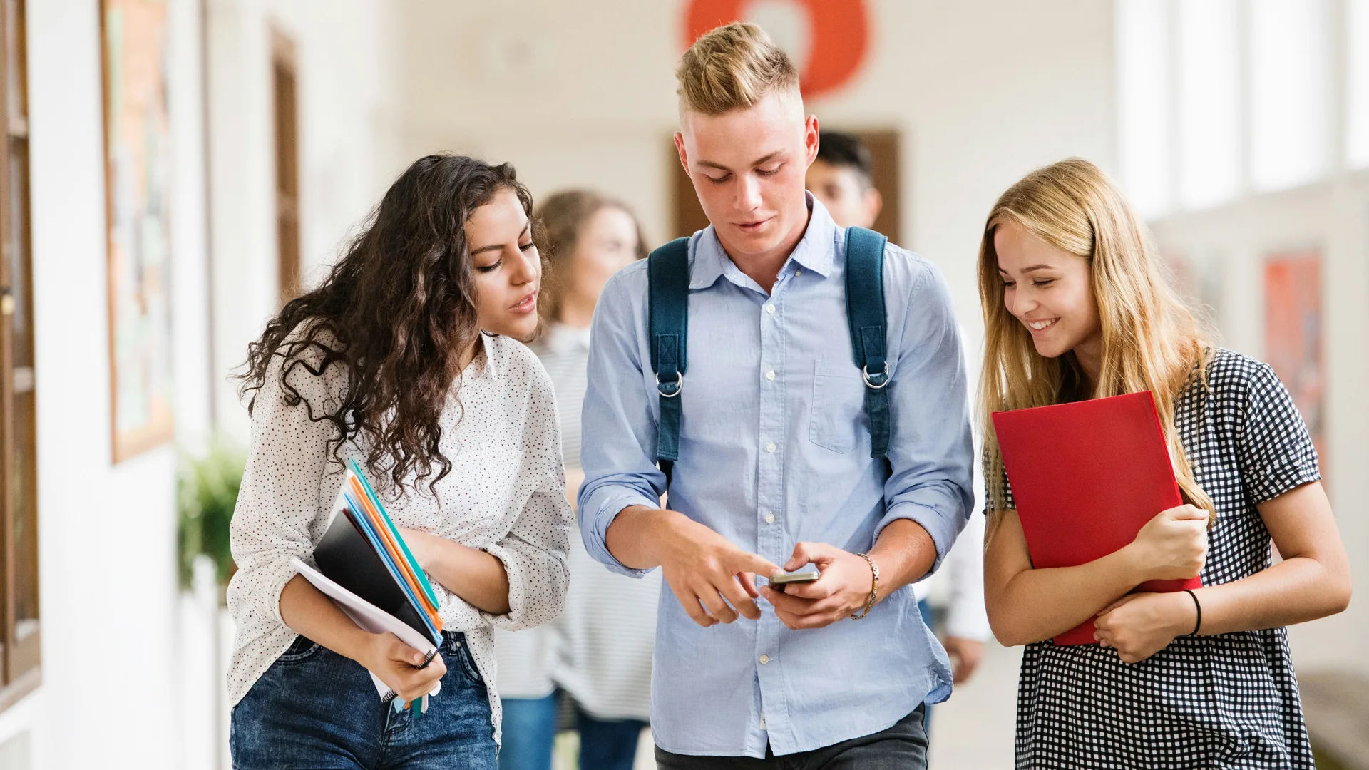 A photo of three teenagers walking through a school hall looking at their phone.