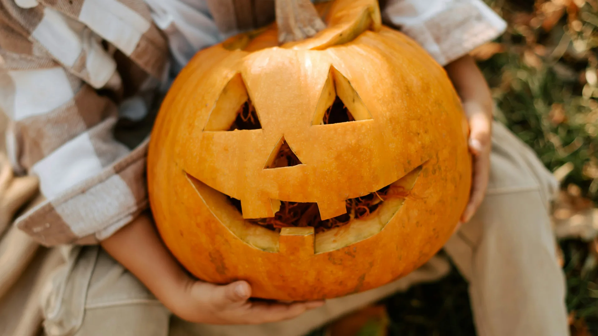 A photo of a pumpkin with a classic triangle eyes and nose carving anf grin with two front teeth. It is being held by someone in a plaid shirt.