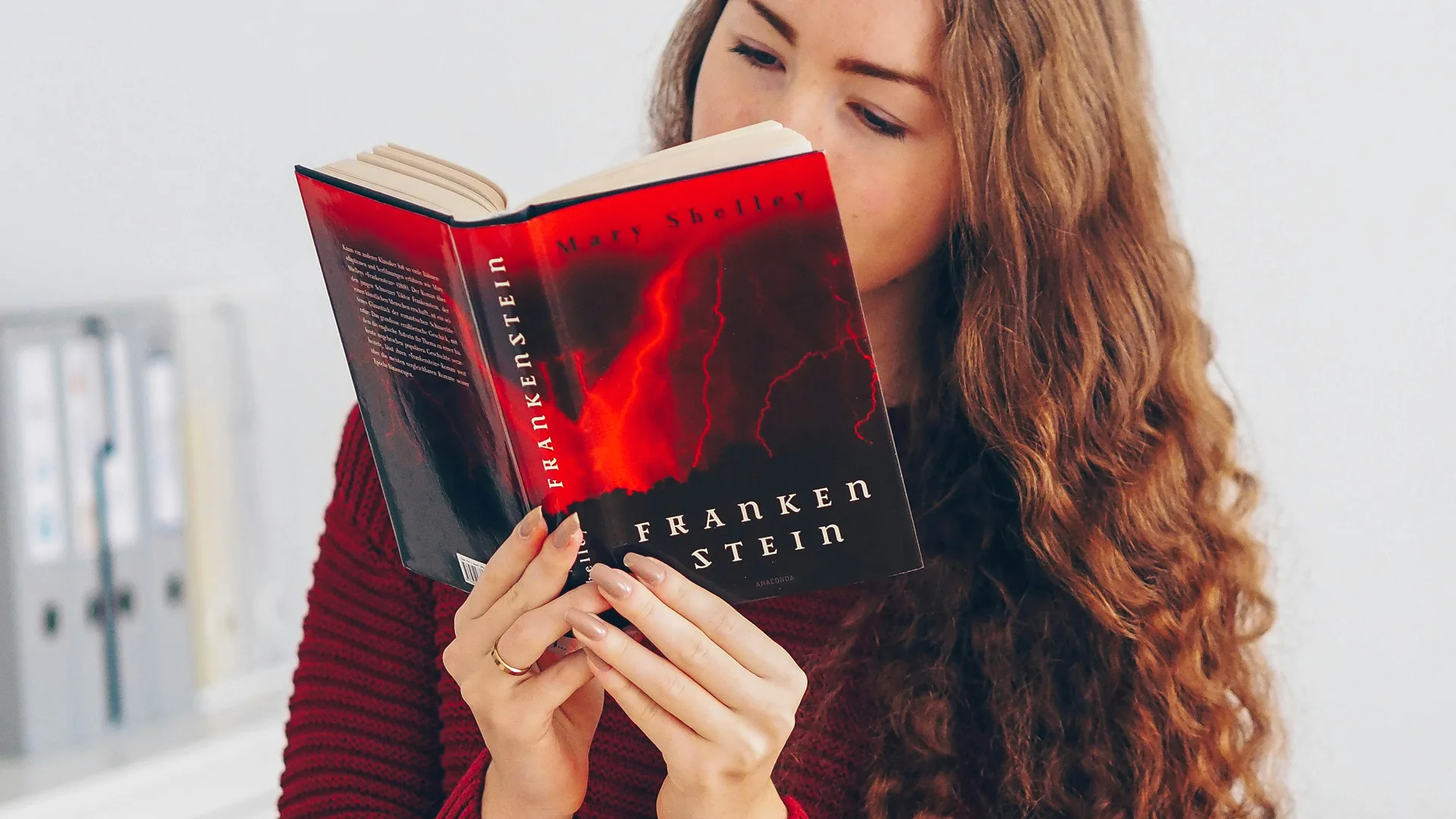 A photo of a woman reading a book of Frankenstein with a red and black cover.