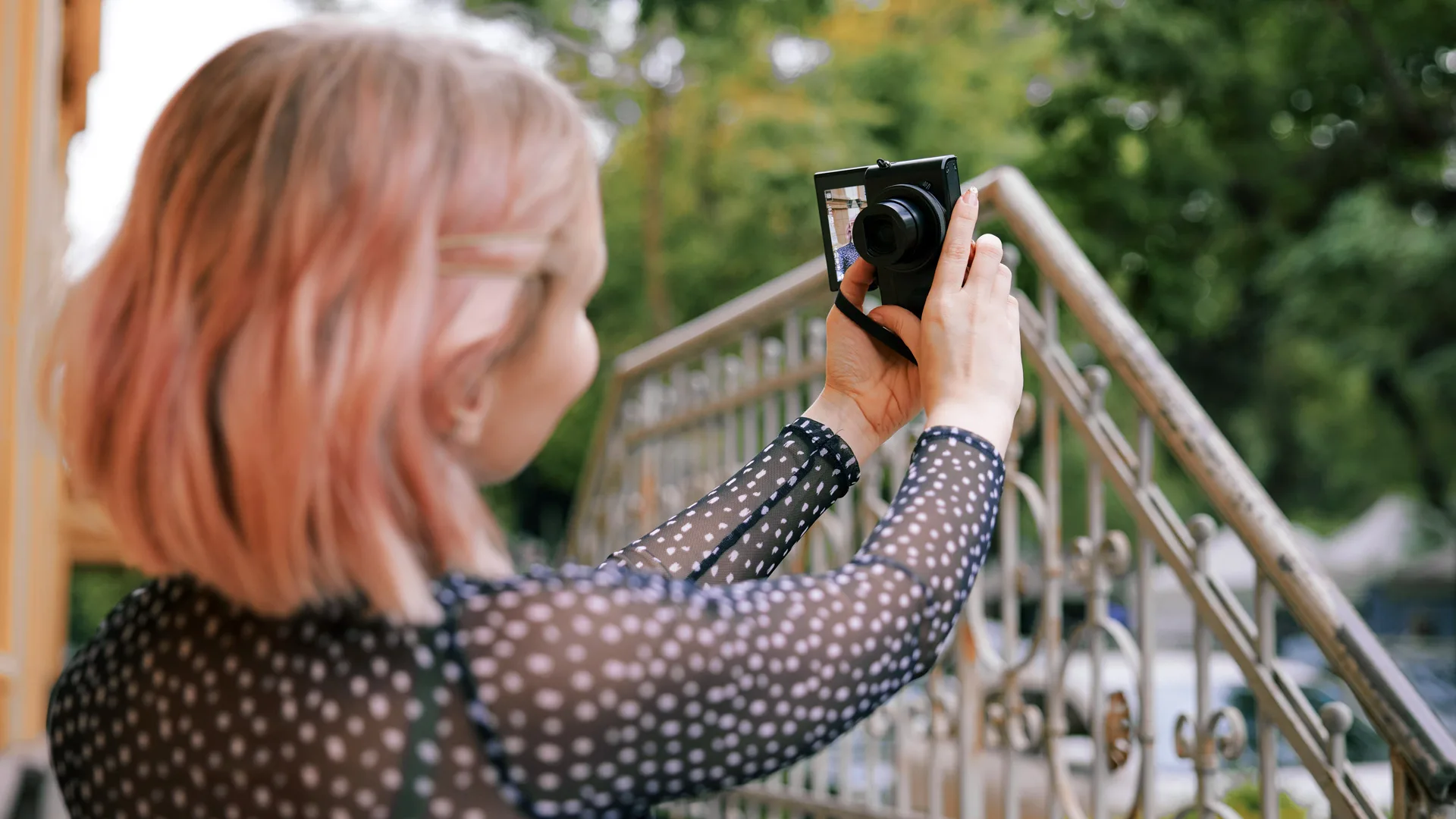 A photo of a woman holding a compact digital camera taking a selfie. She has pink hair and is stood on a bridge with trees in the background.