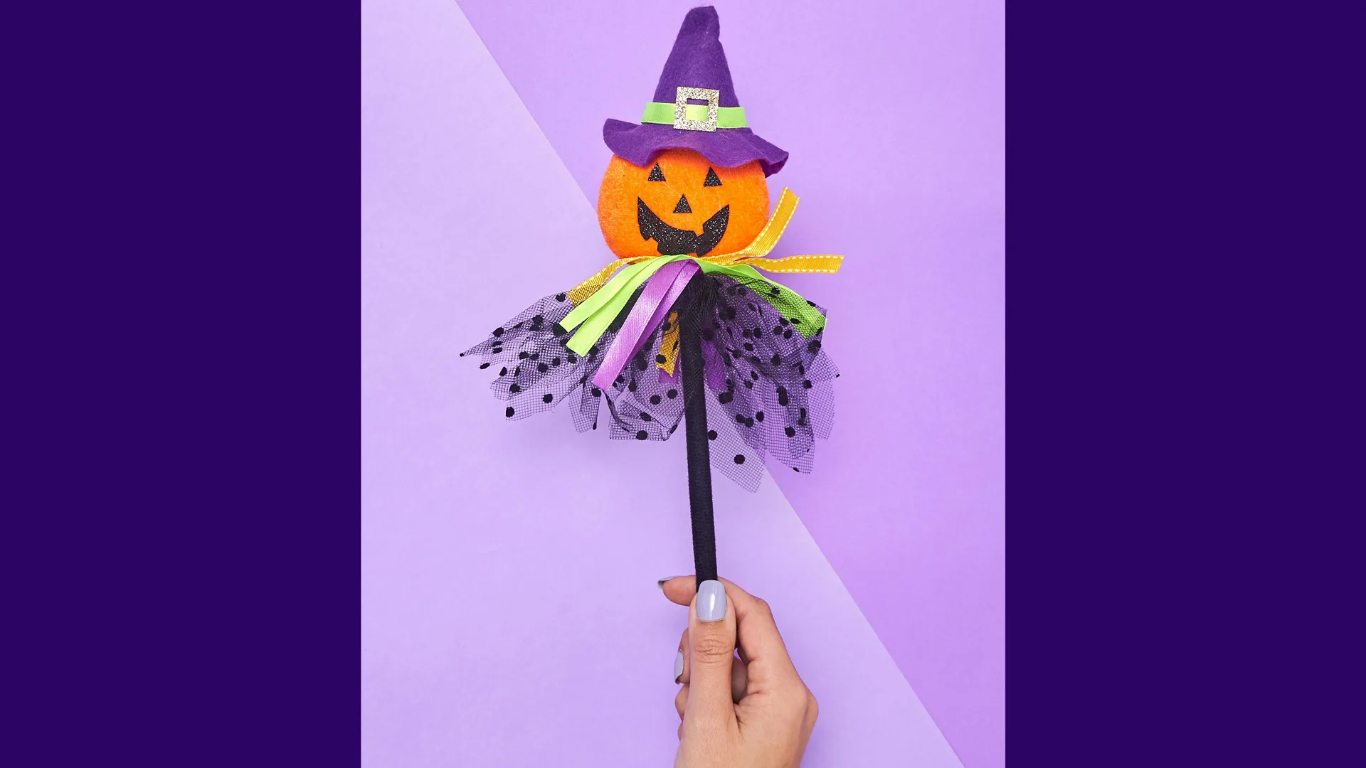 A photo of a pumpkin witch wand craft with a happy orange pumpkin head and purple witch hat with netting and ribbons as a dress, on a black stick held by a hand against a purple background.