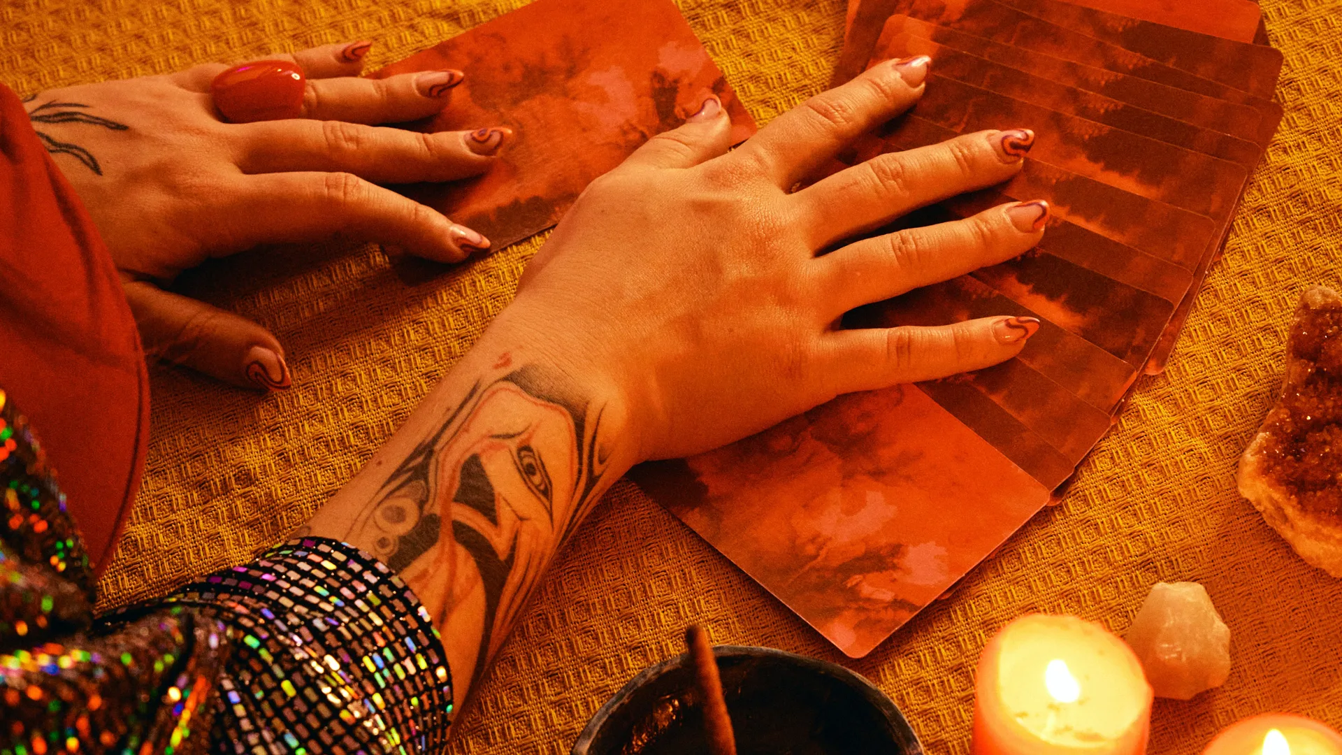 A photo of a hand with tattoos on the arm, spreading out some cards in the orange glow of a burning candle next to it.