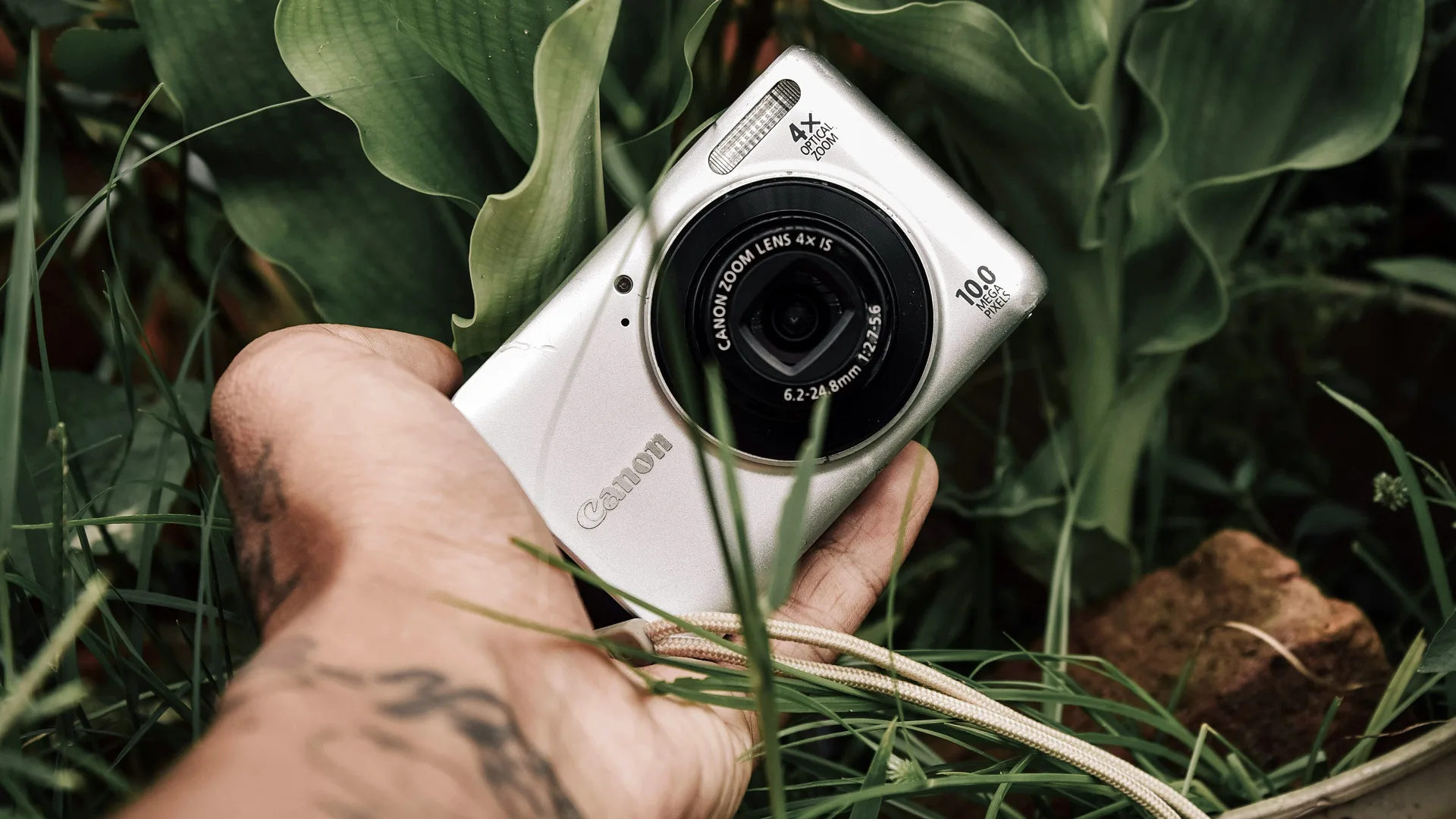 A photo of a canon silver digital camera being held by someone in some grass against some plants.