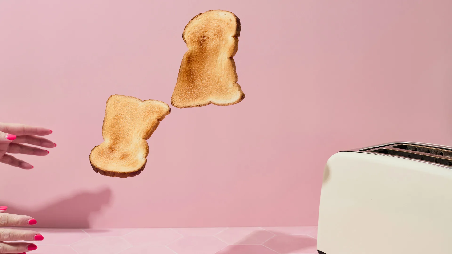 A photo of a white toaster popping two slices of toast mid air with a pair of hands ready to catch them - all against a pink background.