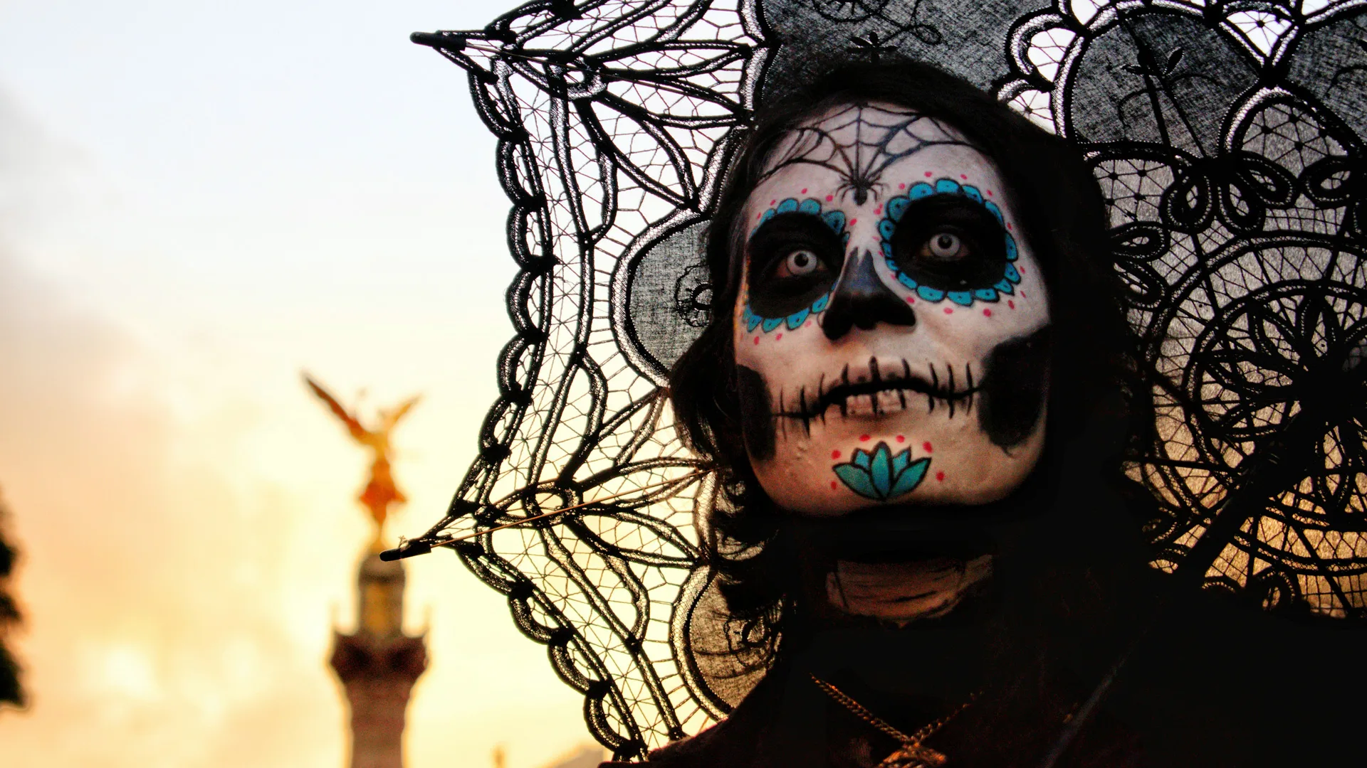 A photo of someone dressed as a Day of the Dead sugar skull with a black lace parasol above their head against a sunset sky.