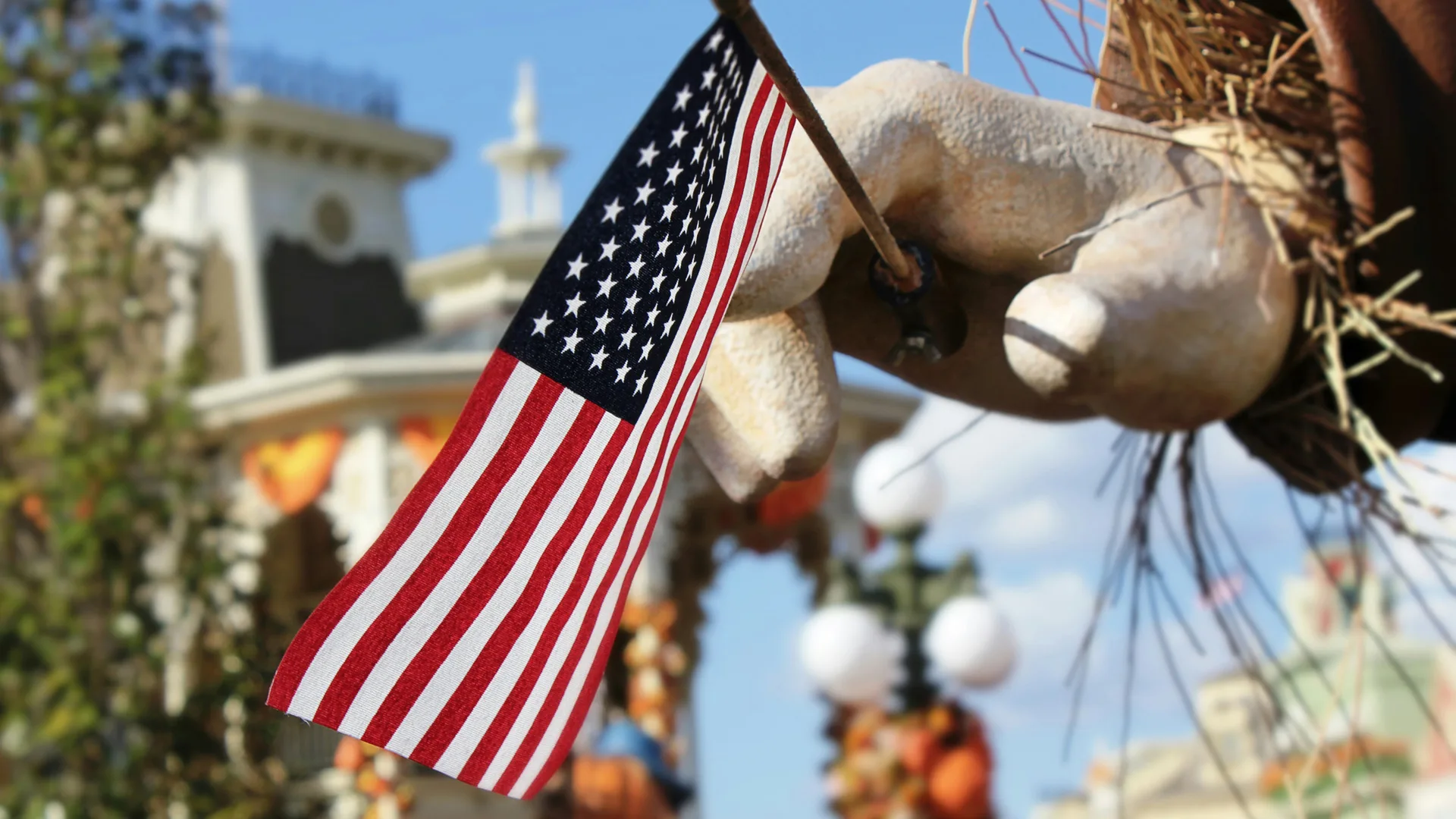 A photo of a wooden hand holding the United States flag in the background are white buildings and Halloween pumpkins