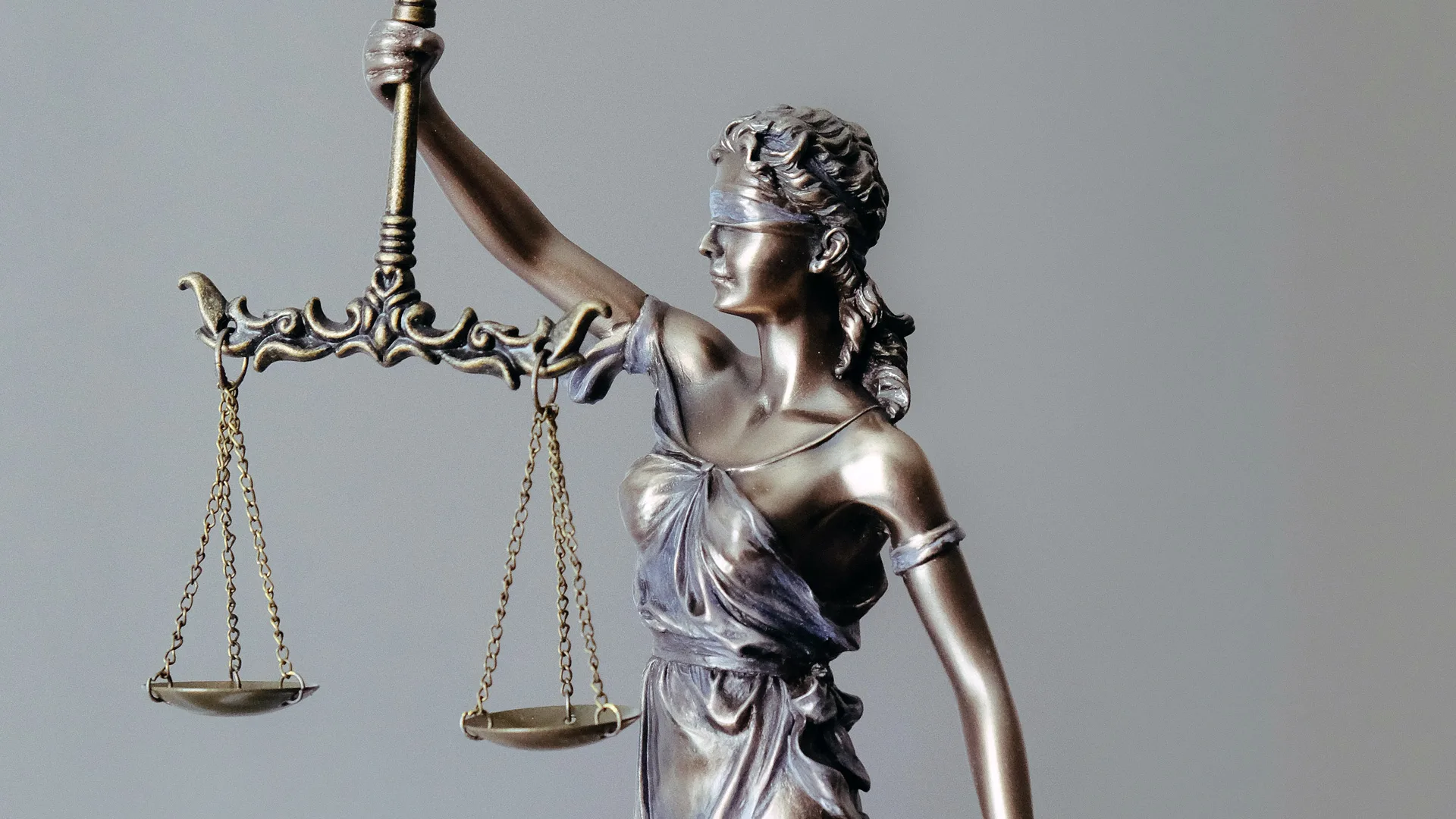 A photo of a statue of a blindfolded lady holding a pair of scales to represent justice in the law. She is stood against a grey background.