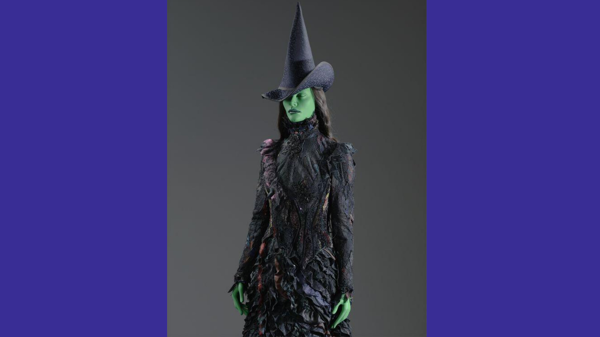 A photo of the Wicked Witch of the West costume in the V&A collection. The costume is a black dress and hat on a green faced mannequin against a grey background with purple borders.