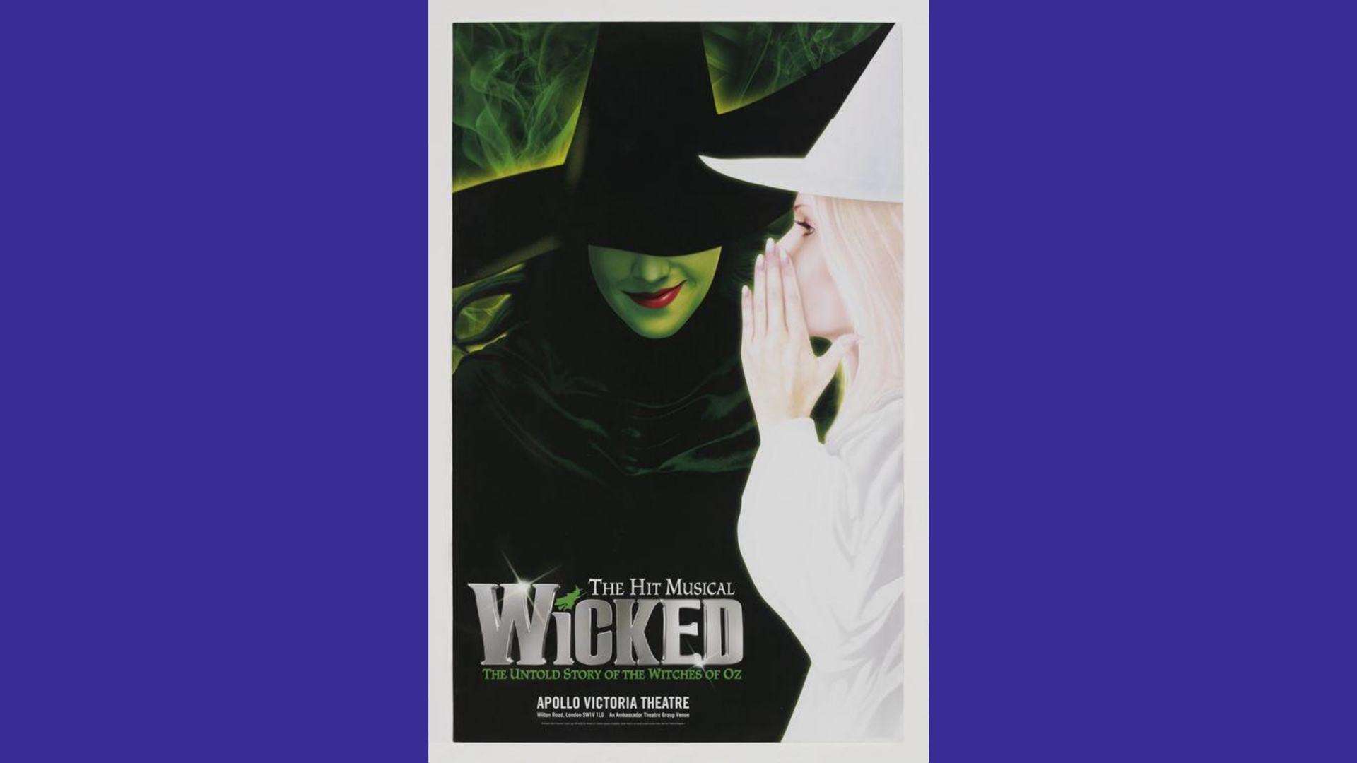 A photo of the Wicked musical poster for the Apollo Theatre in Victoria, London showing Glinda wearing all white whispering into Elphaba's ear who is smiling underneath her large witch hat which is covering her eyes. The picture has a purple border.