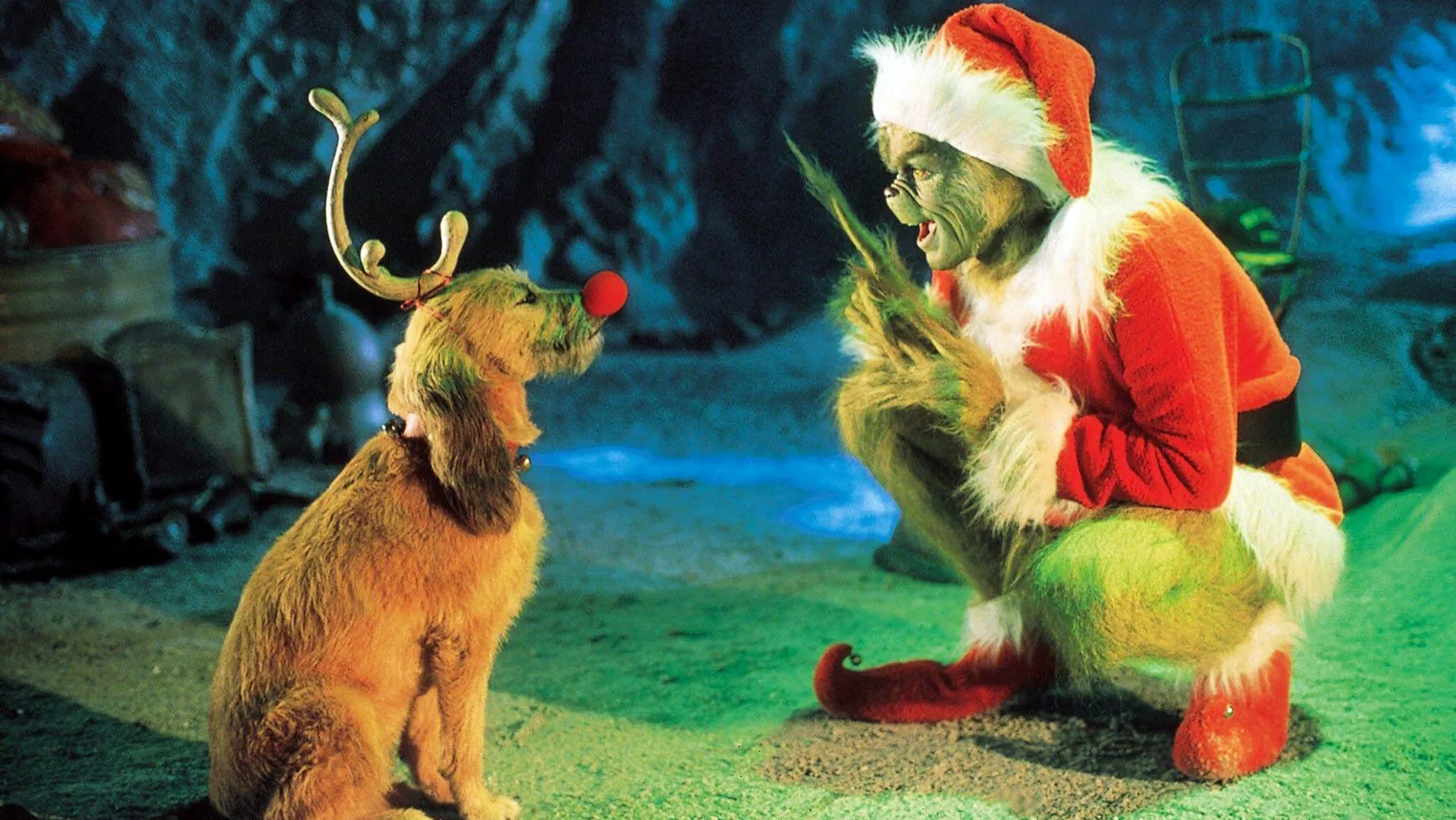 A movie still of The Grinch showing the Grinch wearing his Santa outfit kneeling down looking at his dog who is wearing reindeer antlers and a red nose against a snowy forest background.