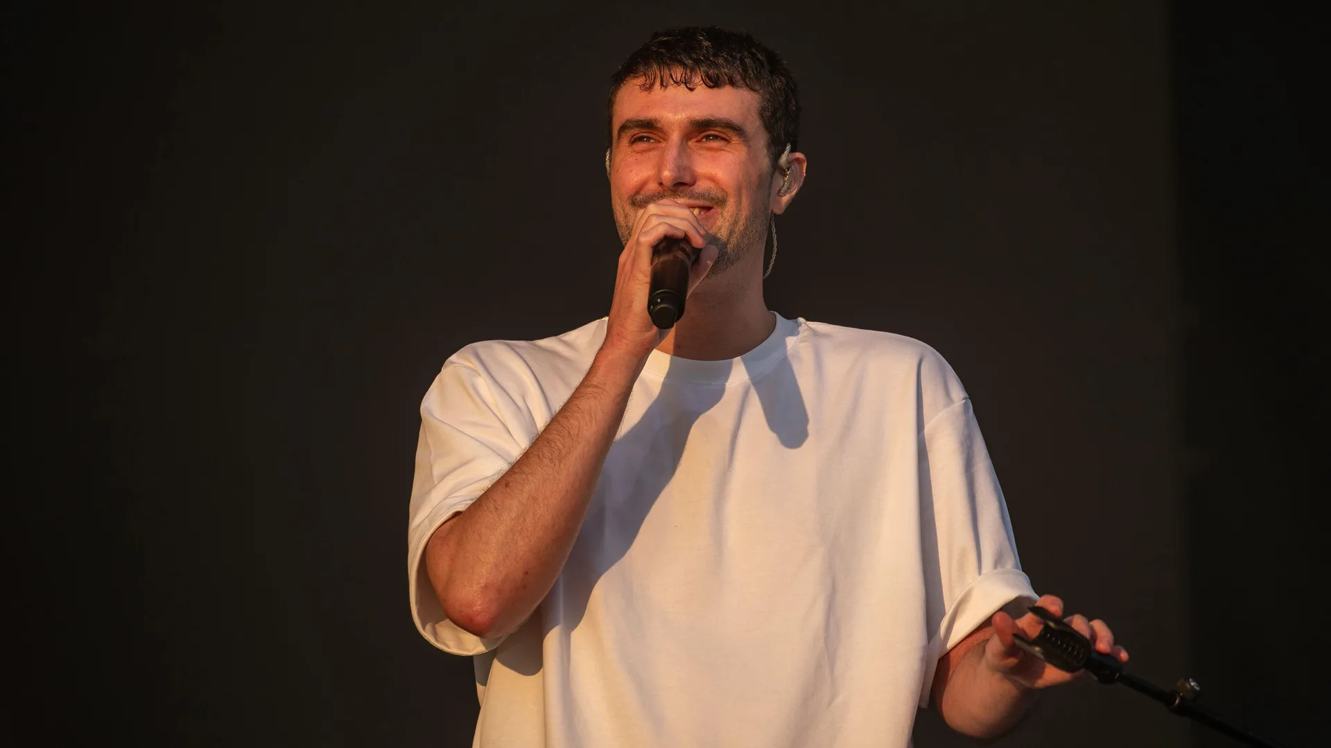 A photo of the musician Fred Again stood on stage smiling with a mic in hand wearing a white tshirt against a black background.