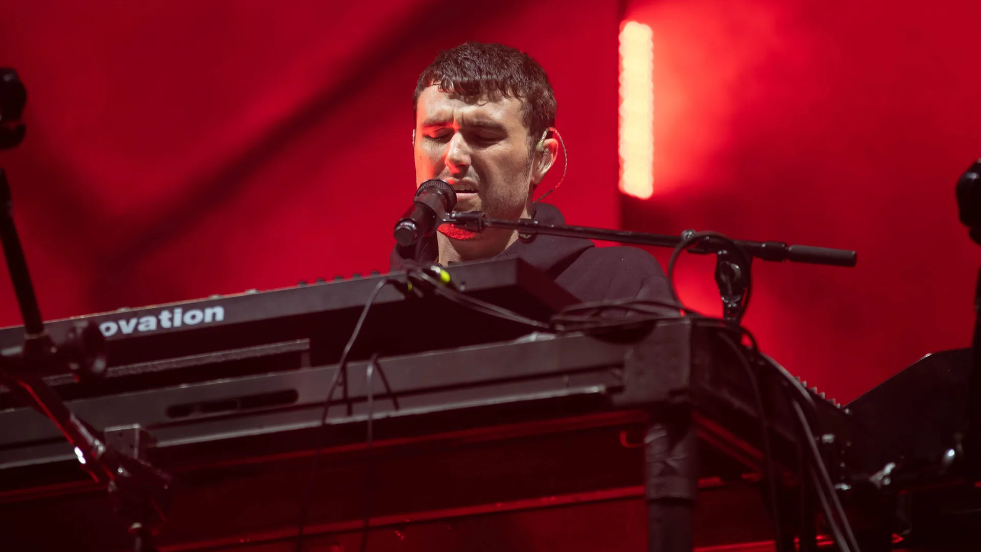 A photo of Fred Again performing on stage against red lights, singing into a mic mounted on a DJ deck.