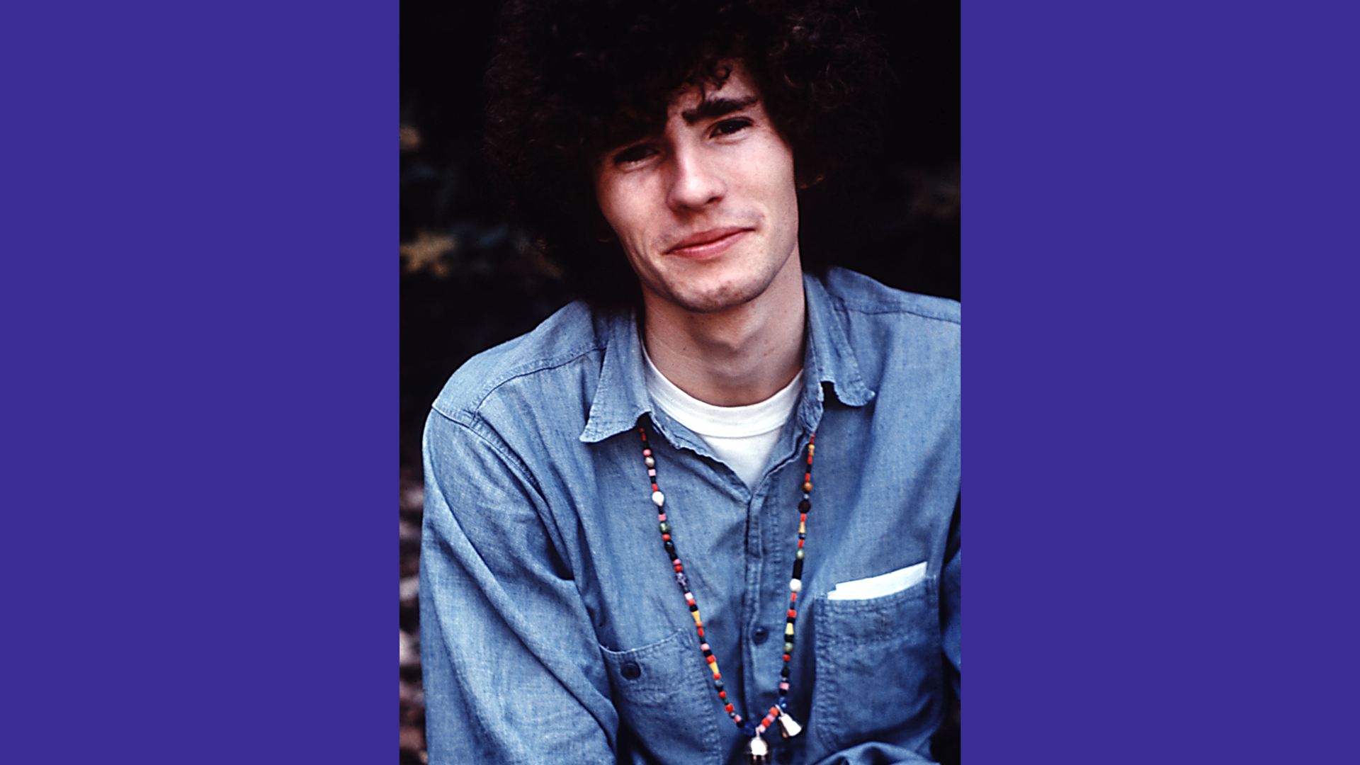 A photo of Tim Buckley the singer, wearing a blue shirt smiling directly at the camera against a black background with purple borders.